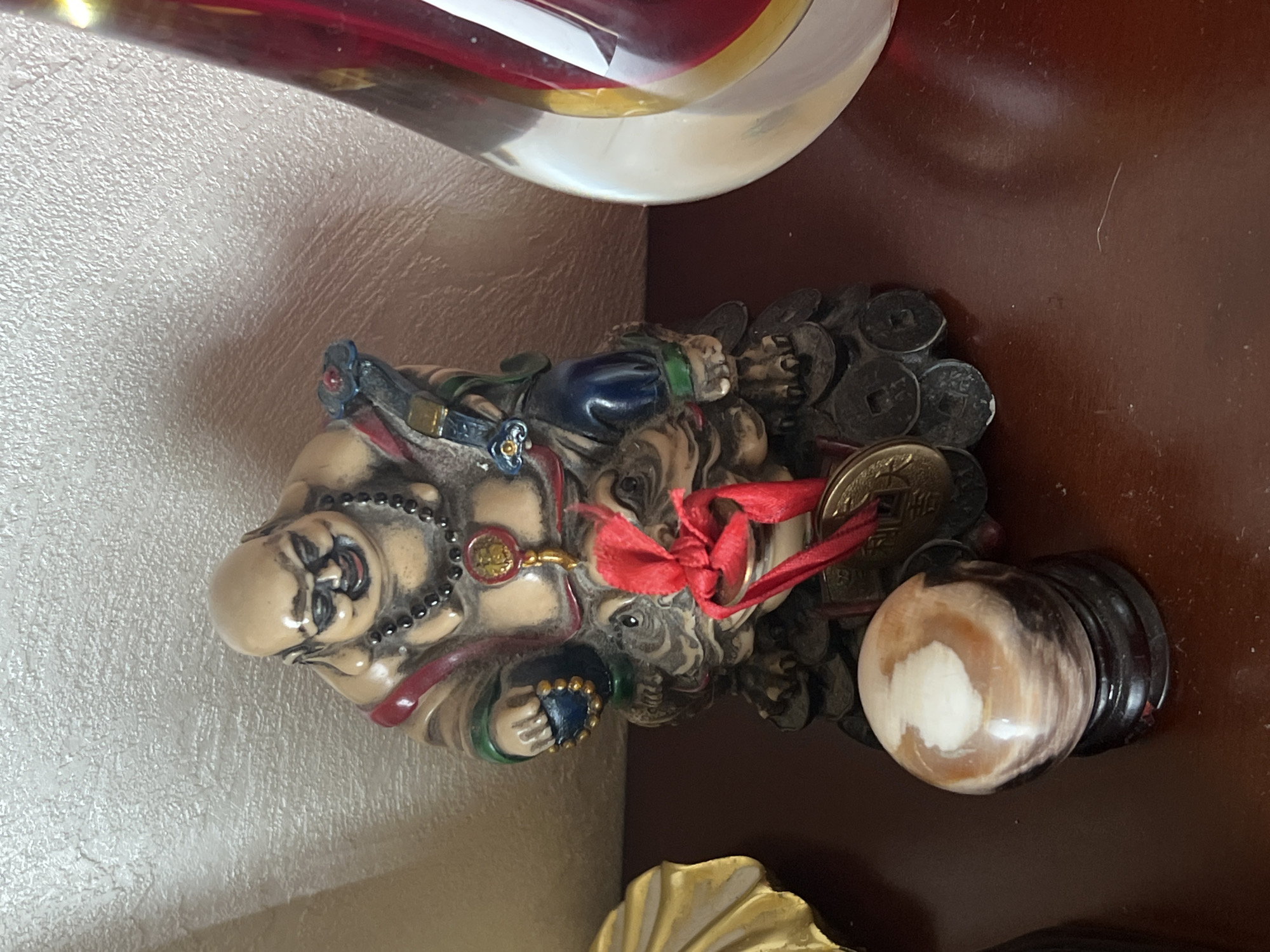 An indoor shot shows a statue of a laughing Buddha, a decorative sphere, and part of a red and clear glass object on a dark wooden surface. The Buddha statue is seated on a mythological creature and is adorned with a necklace, bracelets, and rings. A red ribbon is tied around the statue, with coins attached. To the left of the Buddha statue is a decorative sphere with swirling patterns of brown, white, and cream. To the right, a tall, clear glass object with red and yellow accents is partially visible. The background is a textured, light-colored wall. The image is a close-up, focusing on the arrangement of these objects.