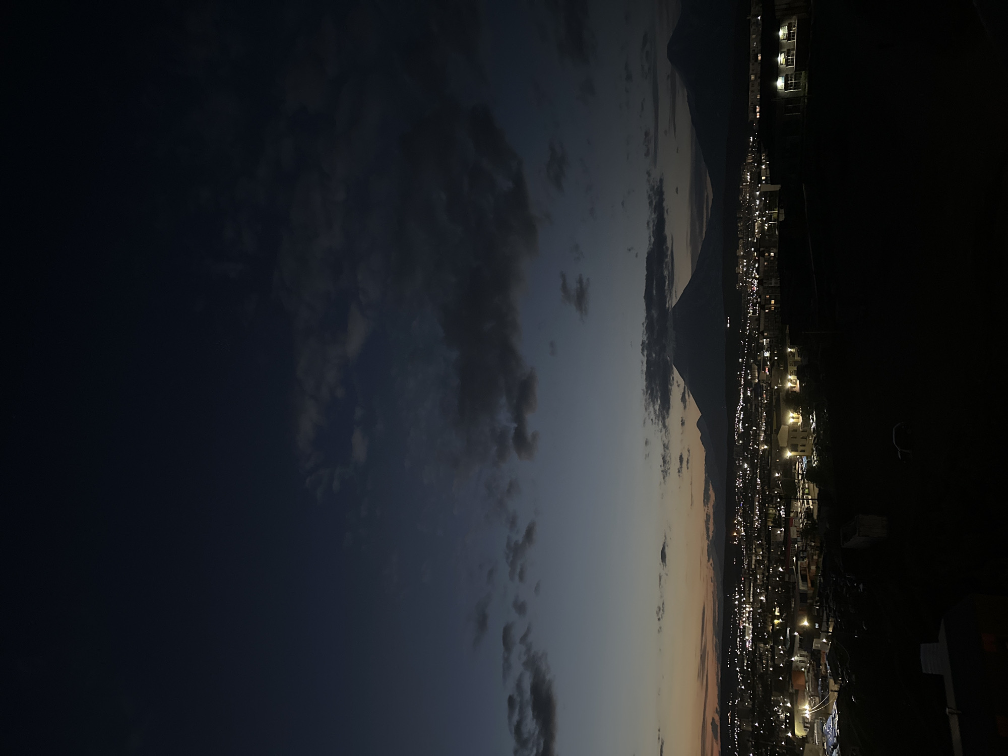 The image depicts a nighttime panoramic view of Petropavlovsk-Kamchatsky, Russia. The sky is a deep twilight blue, with scattered dark clouds and a faint glow of orange and yellow on the horizon, suggesting either the very end of sunset or the very beginning of sunrise. Dominating the background are the silhouettes of two large volcanic cones, which are characteristic of the region. In the middle ground, the city lights are visible, forming a sprawling network of yellow and white points against the dark landscape. The city appears to be built on a gentle slope, with numerous buildings and structures illuminated. The foreground is predominantly dark, with the shapes of hills or elevated terrain covered in shadow. There are no visible people or animals in the frame. The overall atmosphere is one of a quiet, nocturnal urban and natural landscape.