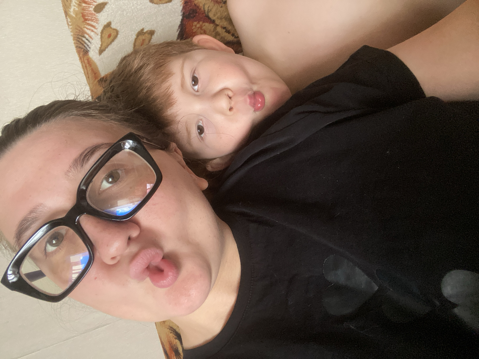 The image shows a close-up selfie of a woman and a young boy. The woman is wearing black-rimmed glasses and a black t-shirt with a heart design. She is puckering her lips. The boy, positioned to her right, is also puckering his lips and looking at the camera. He has reddish-brown hair and fair skin with freckles. They appear to be indoors, possibly on a couch, as a patterned fabric can be seen in the background. The lighting suggests it is daytime. The overall atmosphere is informal and playful. There is no discernible indication of the city of Chaikovsky, Russia in the image.