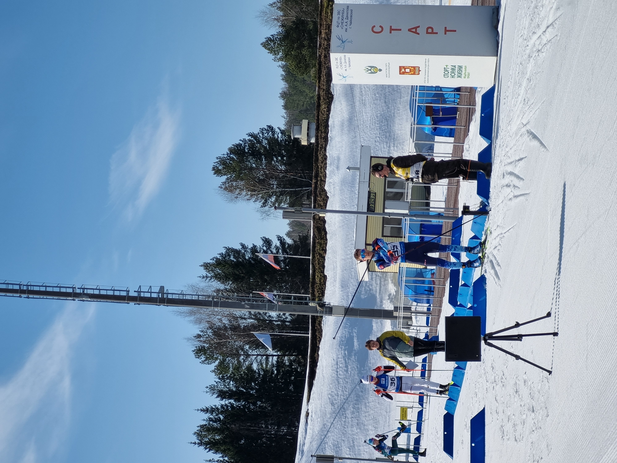The image depicts an outdoor winter sports event at the Federal Center for Winter Sports "Snezhinka" named after A.A. Danilov in Chaykovsky, Perm Krai, Russia. The scene is illuminated by bright daylight under a clear blue sky with a faint wispy cloud.
In the foreground, a black device, possibly a camera or timing sensor, is mounted on a tripod in the center of a groomed, snow-covered surface with parallel ski tracks. To the right of the tripod, blue plastic barriers line the snow.
Five individuals are visible in the mid-ground. From left to right:
1. An athlete in a green and white ski suit, on skis with poles, partially visible.
2. An athlete wearing a white ski suit with blue and red accents, bib number 36, on skis with poles, and carrying a rifle on their back.
3. A person in a yellow and grey jacket and dark pants, holding a clipboard or papers, standing between the two athletes.
4. An athlete in a blue and white patterned ski suit, bib number 35, on skis with poles, and carrying a rifle on their back.
5. An individual in a black jacket, dark pants, and black boots, wearing headphones, with a white bib displaying "64" on their left side. This person stands beside a large white vertical sign.
The large white sign on the right side of the frame displays "СТАРТ" (START) vertically. It also features the emblems of the Russian Ministry of Sport and Perm Krai, and text identifying the venue and the "Sport – Norm of Life" federal project. Behind this sign and the individuals are blue tent-like structures.
In the background, a light-colored building is visible with a digital clock displaying "13:34:39". A tall metal utility pole with ladder rungs extends vertically through the center-left of the frame. Further back, a snow-covered embankment rises, topped with dense coniferous trees and some bare deciduous trees. Three flags are flying on poles on the embankment: the Russian flag, the Perm Krai flag, and a light blue flag.