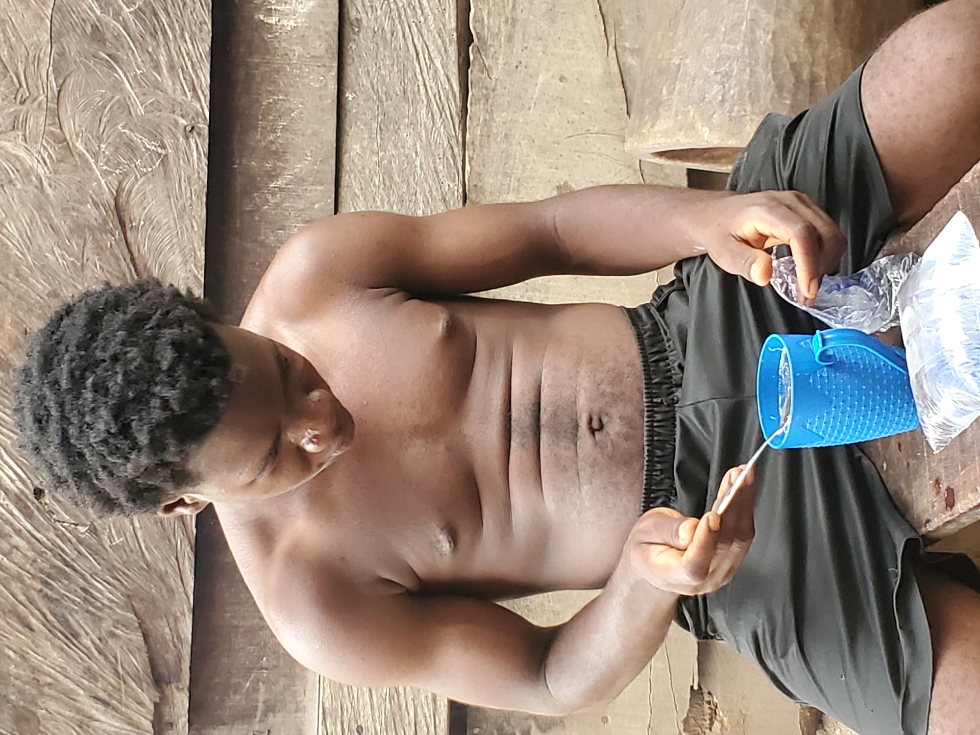 A young, dark-skinned man with short, curly black hair is depicted sitting on a rustic wooden surface. He is shirtless and wears dark-colored shorts. The background and the surface he is on are made of rough wooden planks, exhibiting natural grain and some wear, indicative of a simple, possibly rural or utilitarian setting in Mararaba, Nigeria.
The man is looking downwards with a focused expression, engaged in an activity involving his hands. In his left hand, he holds a small, thin, light-colored object, possibly a spoon or a stir stick. His right hand is seen manipulating a clear plastic bag, which appears to be either opened or being prepared for use.
Beside him, on the wooden surface, is a blue plastic cup or container with a distinctive textured, possibly perforated, surface. Several other clear plastic bags are also visible, some appearing to contain liquid or small items. The arrangement of these objects suggests the preparation or consumption of food or drink. The scene is illuminated by natural daylight. No discernable text is visible in the image.