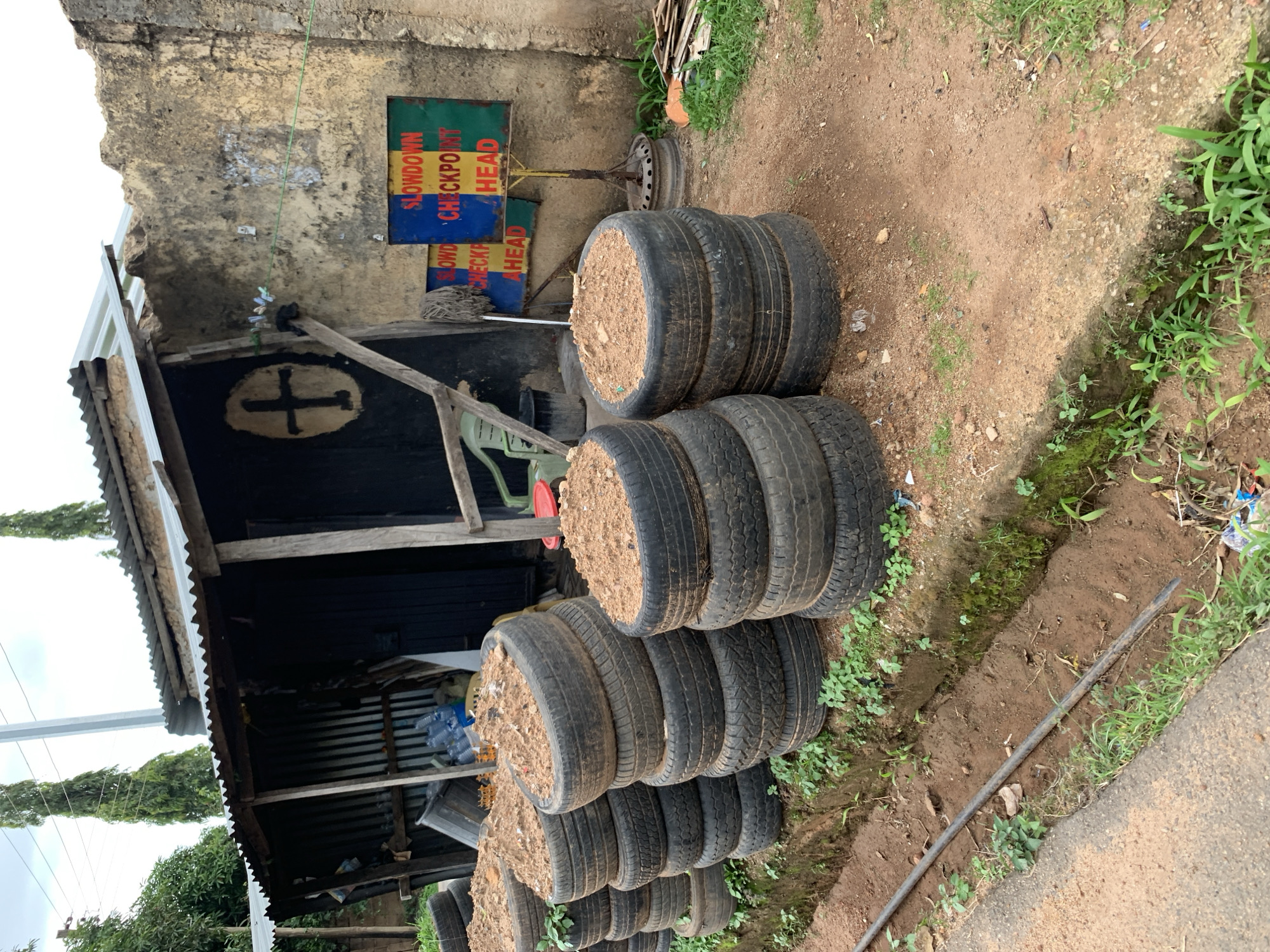 The image depicts a roadside scene in Jos, Nigeria, characterized by a makeshift structure with a corrugated metal roof and walls. In front of this structure, several stacks of tires filled with soil are arranged, suggesting a possible gardening or planting initiative. A sign with colorful blocks of red, yellow, and green is visible, bearing the text "SLOWDOWN CHECKPOINT AHEAD," indicating a potential traffic control point or safety reminder. The surrounding environment is rustic, with dirt ground, sparse vegetation, and the presence of various objects including plastic bottles and a mop. The overall impression is one of a functional, albeit unrefined, community space.
