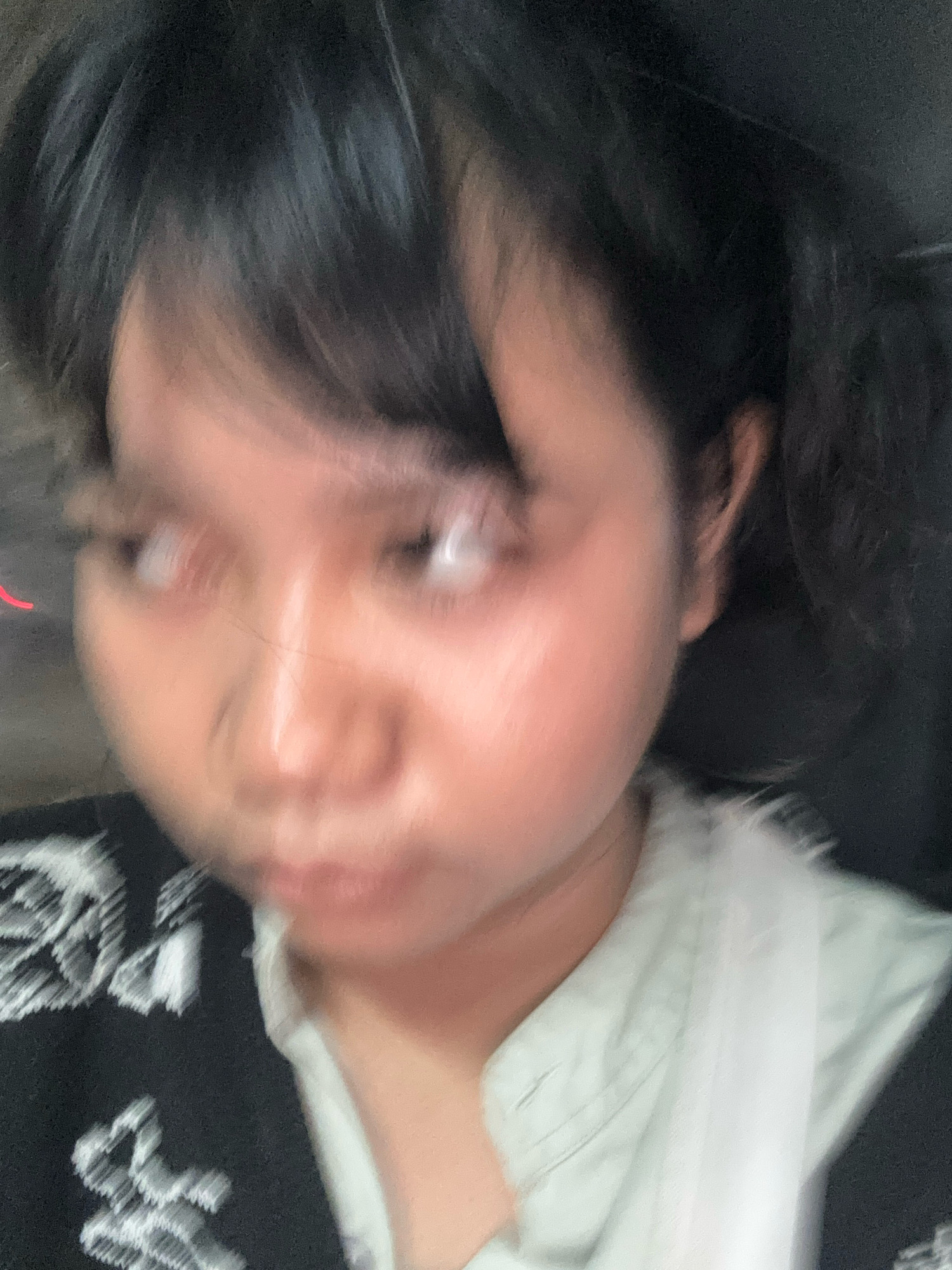 The image is a blurry, close-up selfie of a young Asian individual. The person has dark hair styled with bangs, framing a round face. Their skin appears fair with a rosy blush on the cheeks. The eyes are open and looking upwards. The individual is wearing a black garment with a white embroidered pattern, layered over a light-colored collared shirt. The background is dark and indistinct, suggesting an indoor setting. The image quality is poor due to motion blur, making precise details difficult to ascertain. There is no discernible location in North Jakarta, Indonesia, depicted in this image.