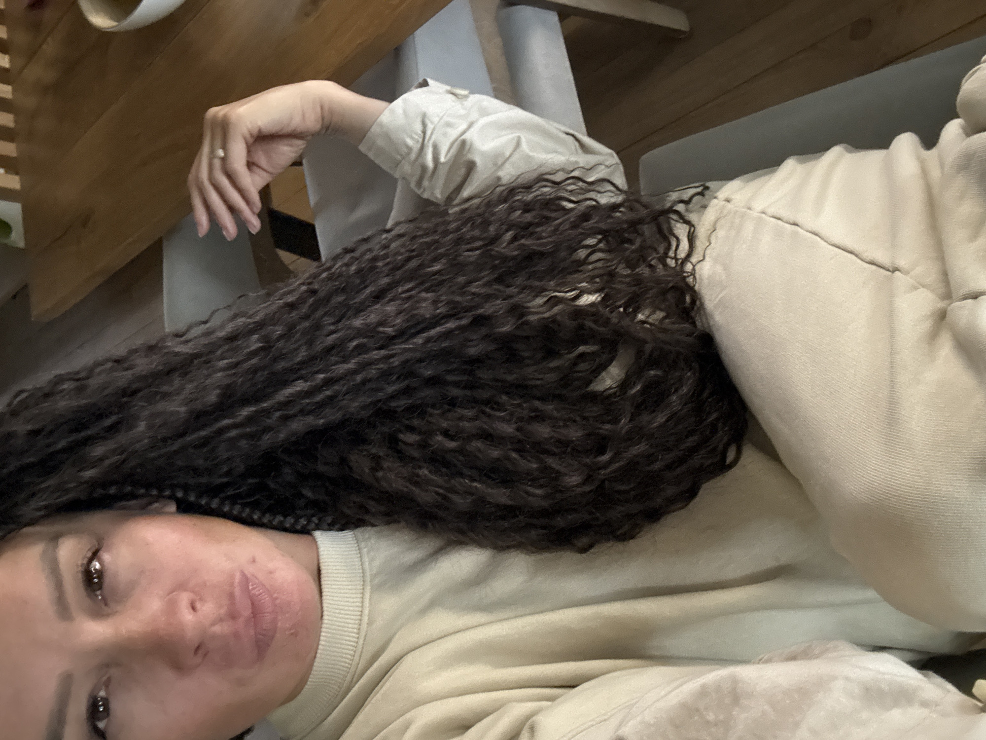 This indoor image, with a provided context of Chaikovsky, Russia, captures a person lying down from a slightly elevated perspective. The main subject is an individual with long, dark, and densely curly or braided hair, which cascades across their chest and arm.
The person's face is visible in the lower left of the frame, looking towards the camera with a neutral and relaxed expression. They have brown eyes. They are wearing a light-colored, possibly beige or off-white, long-sleeved sweatshirt, and what appears to be matching casual pants on the right side of the image, featuring a stitched seam down the leg. A hand, likely belonging to the subject, is seen in the upper left corner, resting on a wooden surface, adorned with a simple ring that has a small, light-colored stone or pearl.
The background shows elements of a domestic setting, including wooden furniture, possibly a table or a headboard, and light-toned support structures or walls. The lighting is soft and consistent, suggesting an indoor environment without harsh shadows. The overall activity depicted is one of relaxation or posing for a casual self-portrait. There is no visible text in the image.