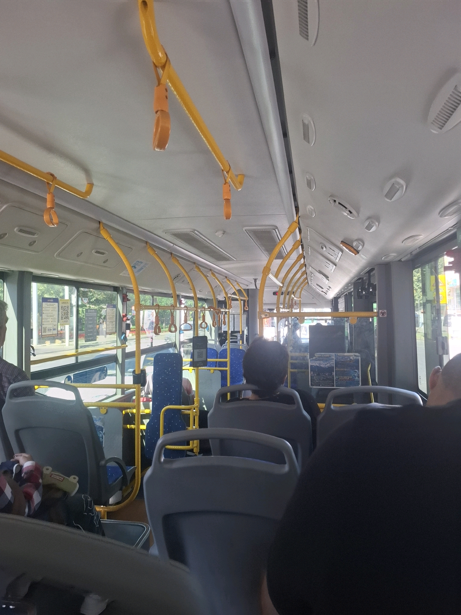 The image depicts the interior of a bus, as seen from a passenger's perspective, looking forward. Yellow handrails and orange hand grips are prominently featured along the ceiling and sides of the bus. Several grey passenger seats are visible, some occupied by individuals. Outside the bus windows, a city street scene with trees and buildings can be observed, suggesting an urban environment. The presence of a QR code on a poster inside the bus, along with Cyrillic text on other signage, indicates the location is likely in a Russian-speaking region, consistent with Almaty, Kazakhstan. The overall lighting suggests it is daytime.