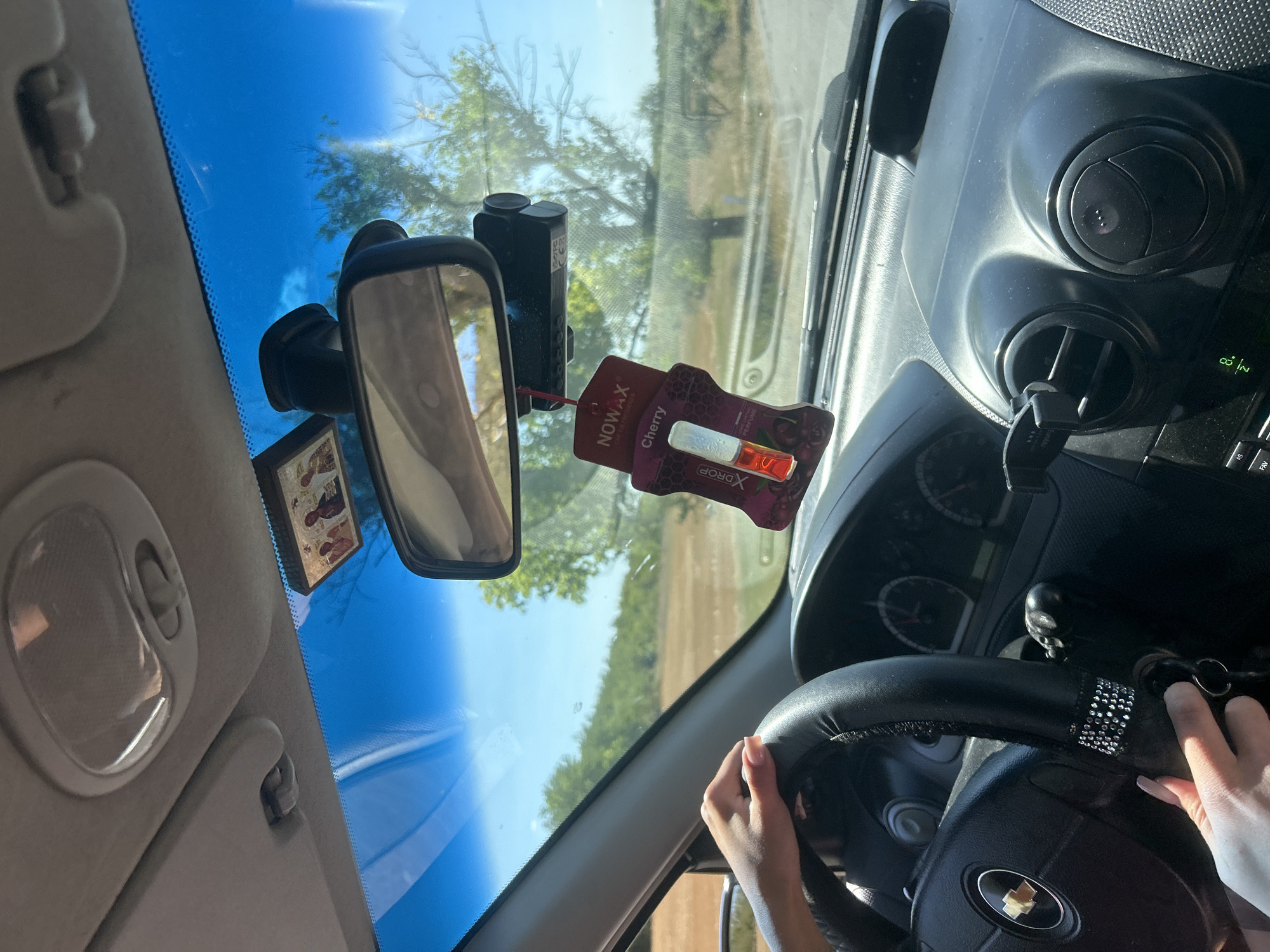 The image depicts the interior of a car, looking forward through the windshield. A person's hands are visible on the steering wheel, which is covered in black leather and decorated with rhinestones. The car is in motion, with a landscape of trees and fields visible through the windshield. A car air freshener hangs from the rearview mirror, along with a dashcam. The dashboard and air vents are also visible. The scene appears to be a sunny day.