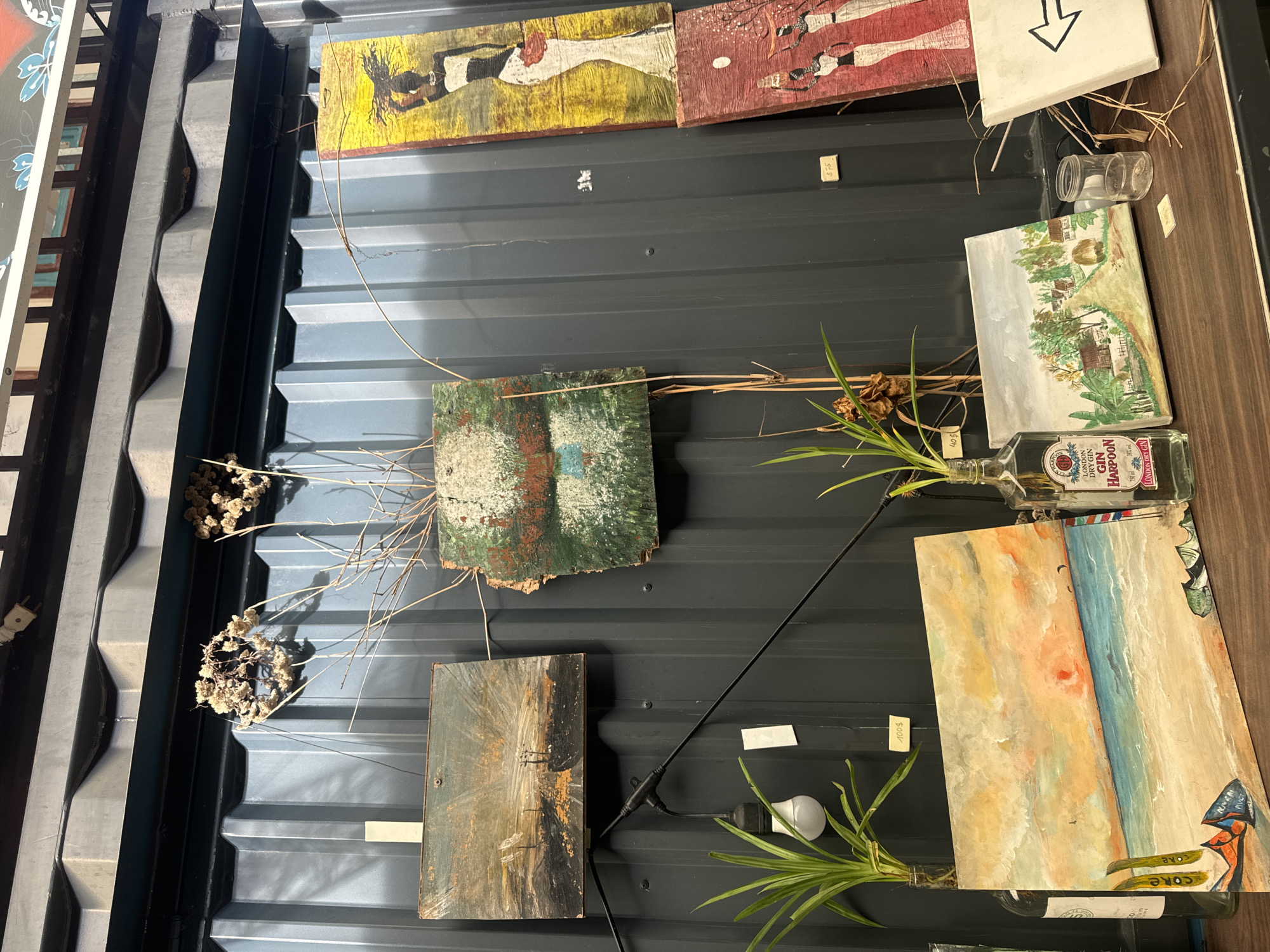 The image depicts an outdoor stall display, likely in Phan Thiết, Vietnam, showcasing various art pieces and decorative items. The backdrop is a dark, corrugated metal wall, possibly part of a shipping container.
Dominating the upper portion of the display are several paintings. To the left, a horizontal painting depicts a seascape with a boat and a stormy sky. To its right, a rough, square piece of wood is painted with abstract green, white, and red textures. Further to the right, two vertical paintings are displayed: the upper one features a silhouetted woman in a white dress against a yellow background, and the lower one shows two figures in profile carrying baskets on their heads against a red backdrop.
Below these, a string of decorative lights with a single exposed bulb hangs. Several small plants in glass bottles are placed on a wooden shelf in the foreground. A bottle of "London Dry Gin Harpoon" is prominently featured, containing a plant.
To the right of the gin bottle, a horizontal painting shows a beach scene with a sunset, sailboats, and surfboards. Next to this, a smaller canvas displays a pastoral landscape with houses and trees. A small glass jar sits between the two paintings. To the far right, a white canvas with a black arrow pointing left is propped up.
Scattered amongst the items are dried branches and flowers, adding to a rustic, bohemian aesthetic. Price tags are visible on some of the artworks, including one for "$75" on the lower vertical painting. The overall scene suggests an art stall or market, with a focus on handmade or vintage items.