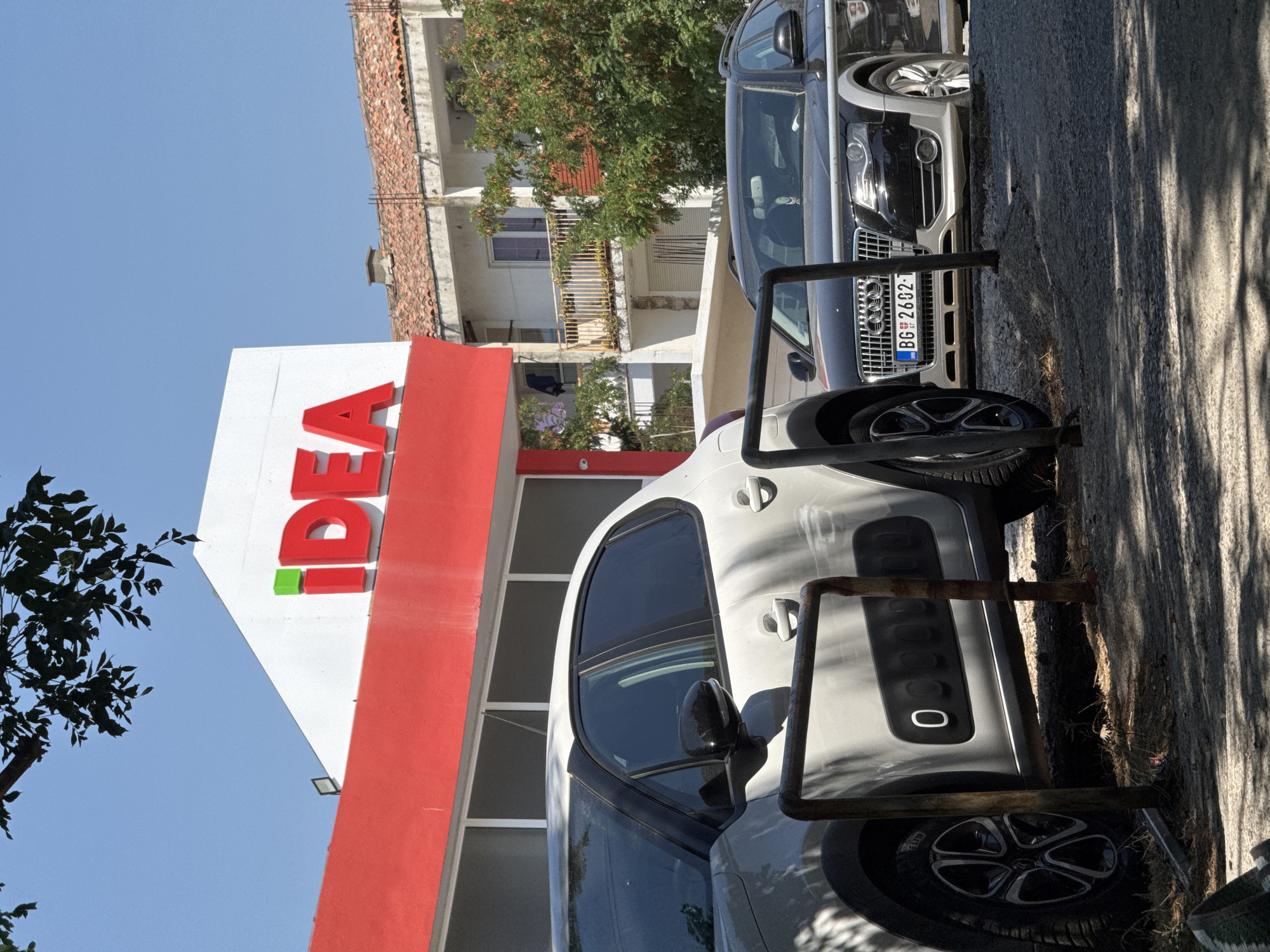 An outdoor, eye-level shot features a supermarket with a red and white facade, displaying the red and green "IDEA" logo. The building is situated in the city of Sutomore, Montenegro. In the foreground, two cars are parked: a white hatchback on the left, and a black Audi wagon on the right. The Audi's license plate is visible, reading "BG 2602-1". A tree with green foliage is visible in the background. The scene is bathed in sunlight, casting shadows on the ground and cars.