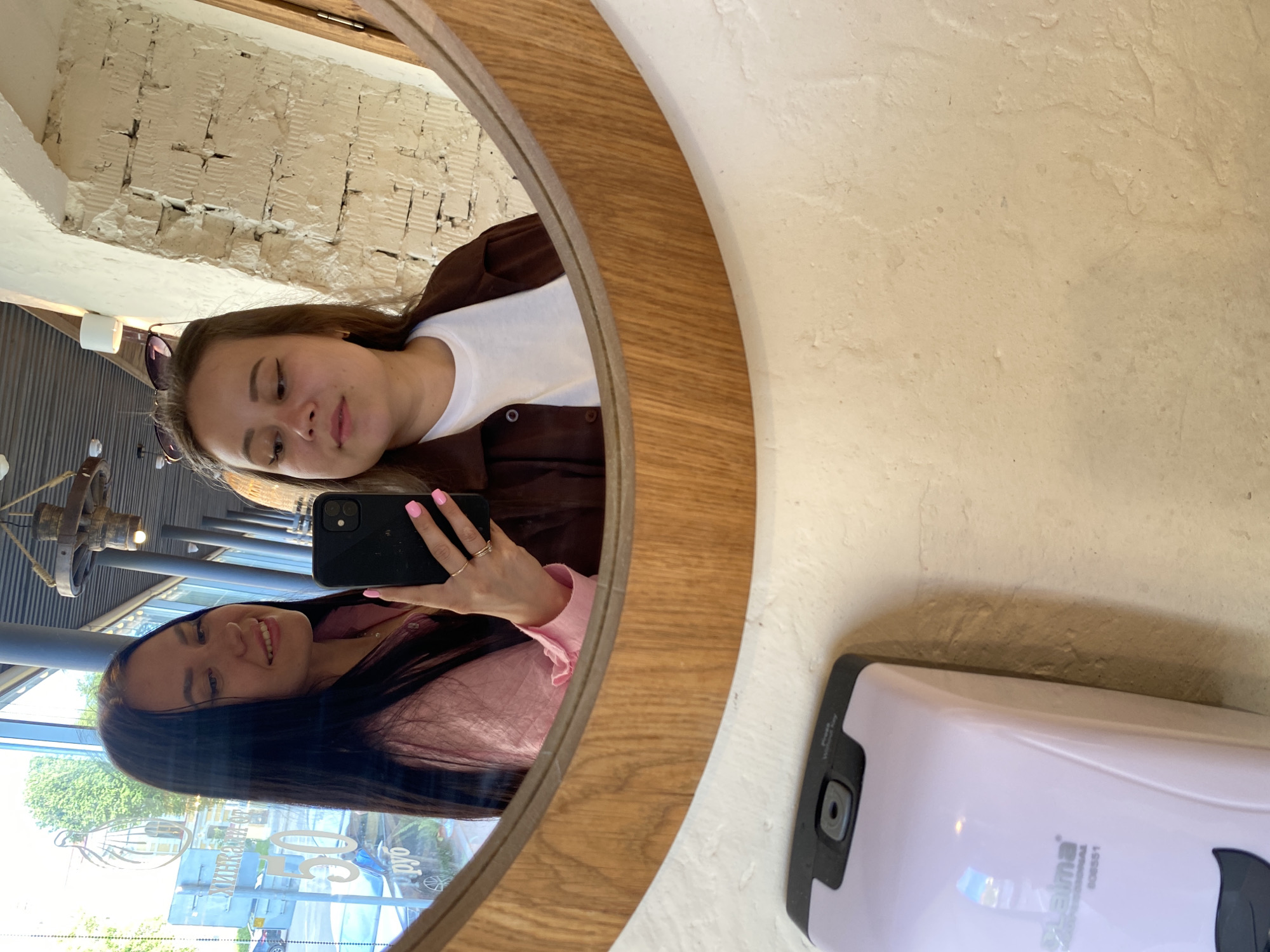 In a mirror at what appears to be a cafe or restaurant in the city of Perm, Russia, two young women are taking a selfie. The woman on the left, with long dark hair, is wearing a pink shirt and has her right hand holding a black smartphone up to take the picture. She is smiling, and her fingernails are painted pink. The woman on the right, with light brown hair, is wearing a white t-shirt and a brown jacket. Sunglasses are perched on her head. The background reflects a large window with signage that includes the number "50" and text in Cyrillic. Below the mirror, a light purple soap dispenser is visible on a textured beige wall. The overall setting suggests an indoor public space with a view to the outside.