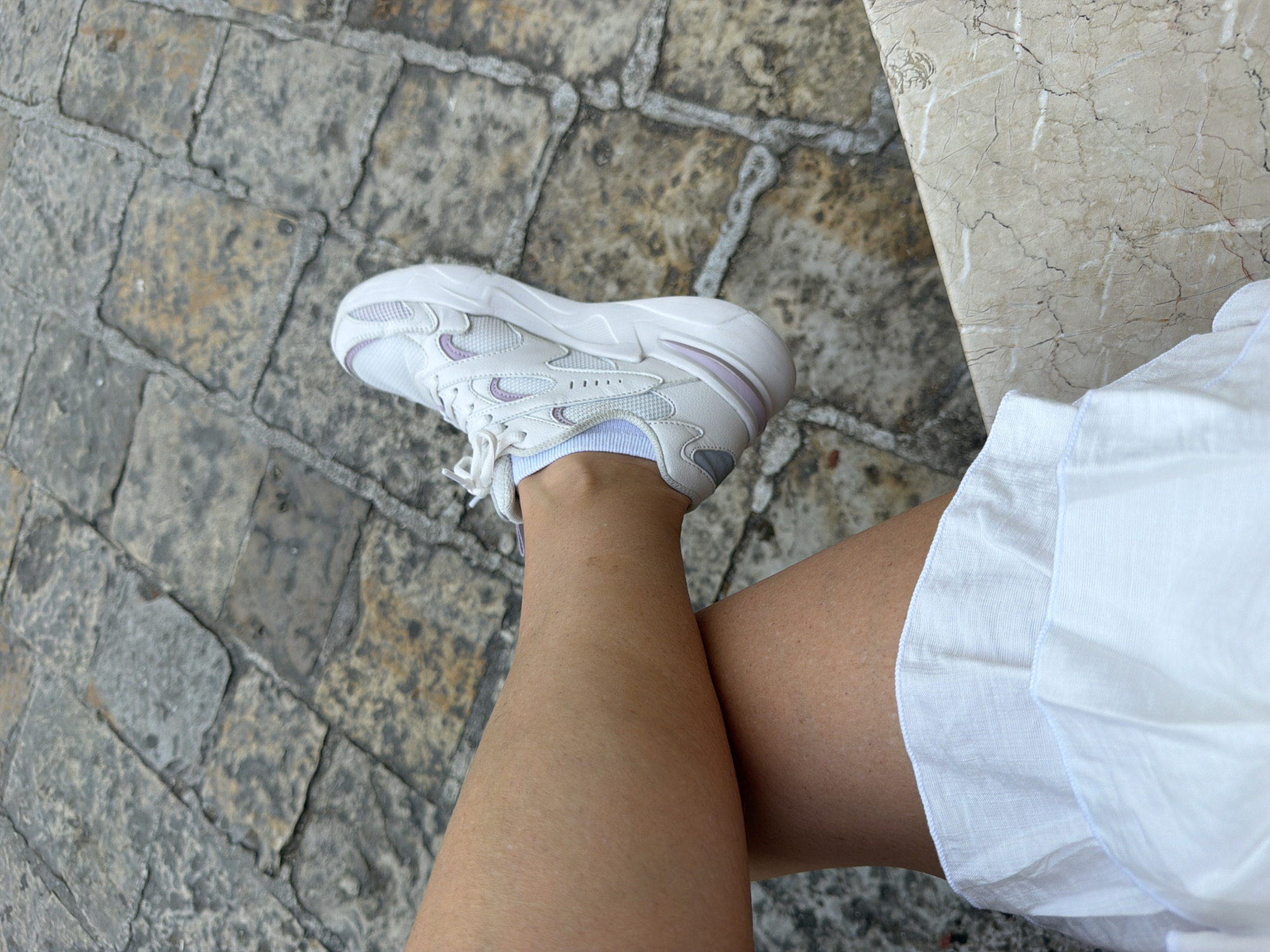 A close-up, high-angle shot shows a person's legs crossed while sitting on a textured marble surface. The individual is wearing white athletic shoes with purple accents and white socks. Their legs are bare, and they are wearing a white ruffled garment around their waist, suggesting a dress or skirt. The background consists of rough, grey cobblestones. The scene appears to be an outdoor seating area in Budva, Montenegro, likely a cafe or restaurant terrace, given the combination of cobblestone paving and marble furnishings.