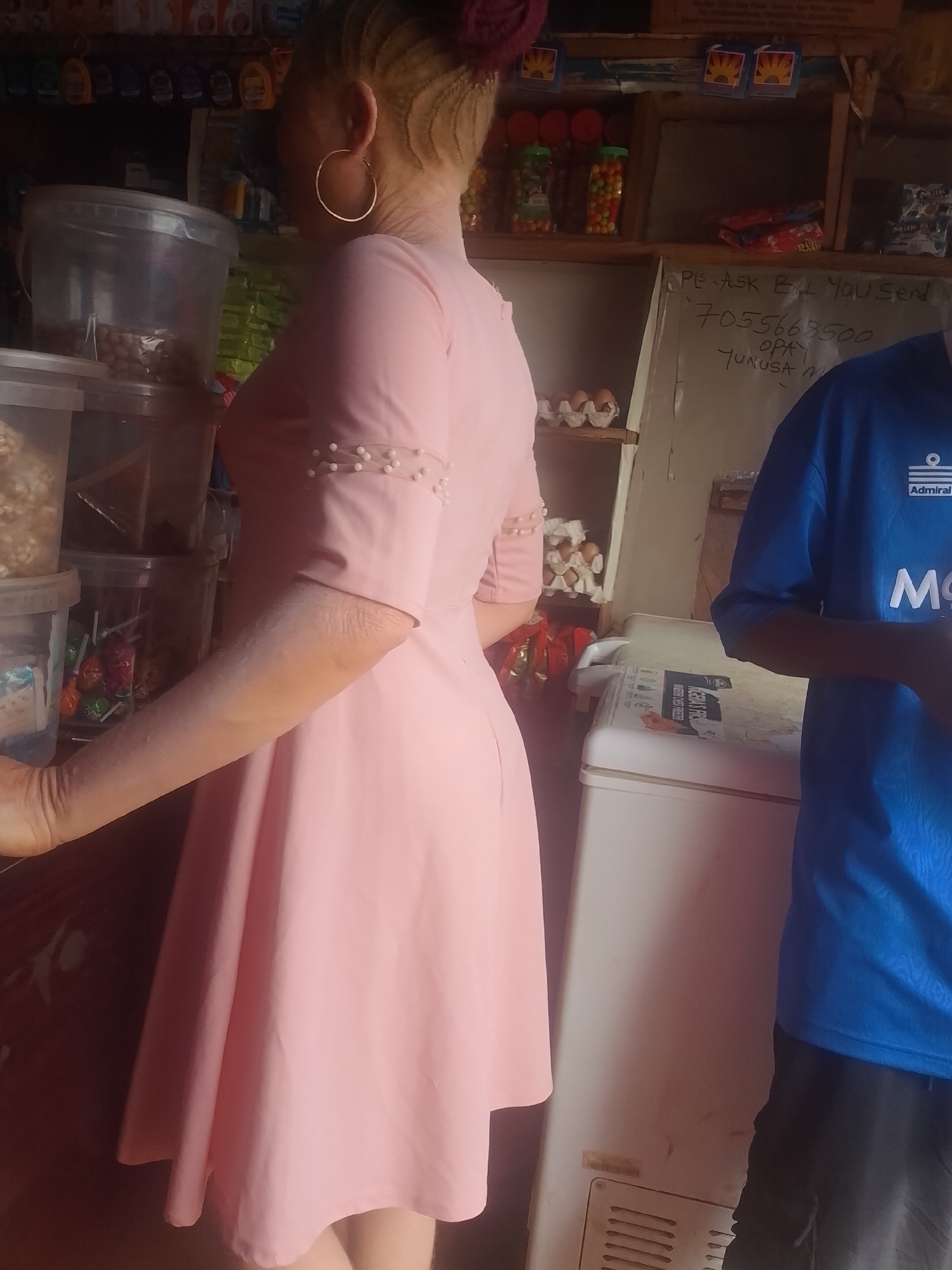 A woman wearing a light pink dress and gold hoop earrings stands at a counter in a small retail shop in Ejule, Nigeria. The shop has shelves stocked with various items, including jars of candy, containers of peanuts, popcorn, and lollipops. A second person, partially visible, wears a blue Admiral brand shirt. A white freezer is adjacent to the counter, and handwritten messages are visible on the wall behind the freezer.
