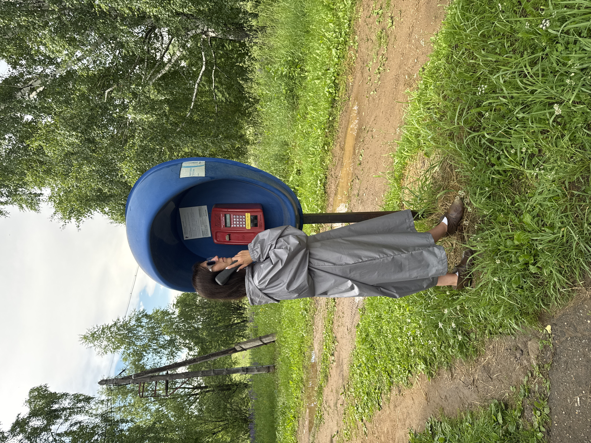 In Вассята, Russia, a woman stands in a grey raincoat by a blue public payphone booth, speaking on the handset. The payphone has a red keypad and display. She is wearing sunglasses and brown shoes. The scene is outdoors, with a dirt road and grass surrounding the payphone. Trees are visible in the background. The sky is partly cloudy. The ground shows signs of recent rain, with puddles on the dirt road. A wooden utility pole with wires is also present.