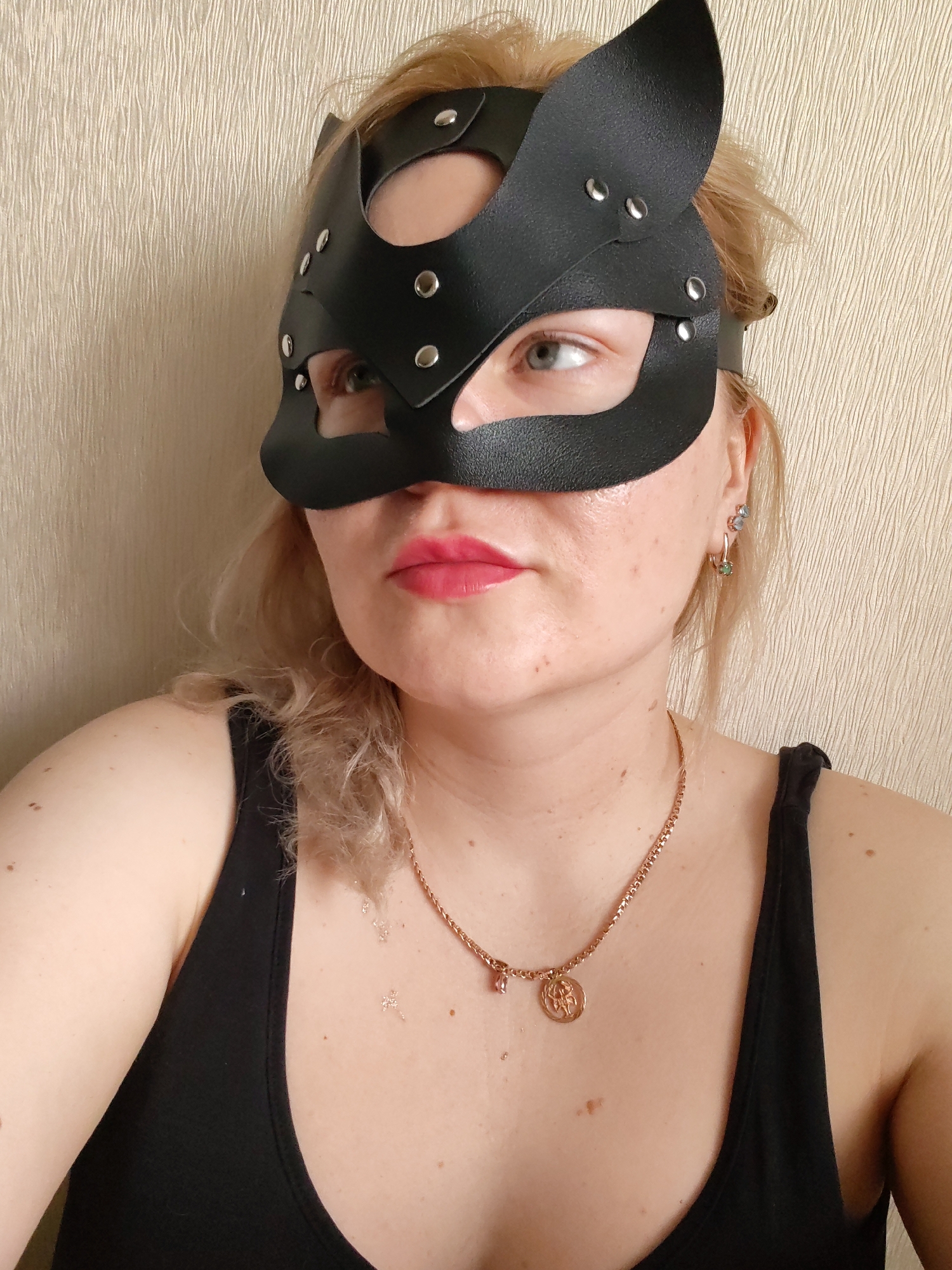A woman wearing a black leather cat mask with metal rivets and a black tank top is posed against a textured beige wall. The woman has blonde hair, light skin with freckles, and pink lipstick. She is also wearing earrings and a gold necklace with a pendant. The woman is looking to the left. No location details are evident in the image.