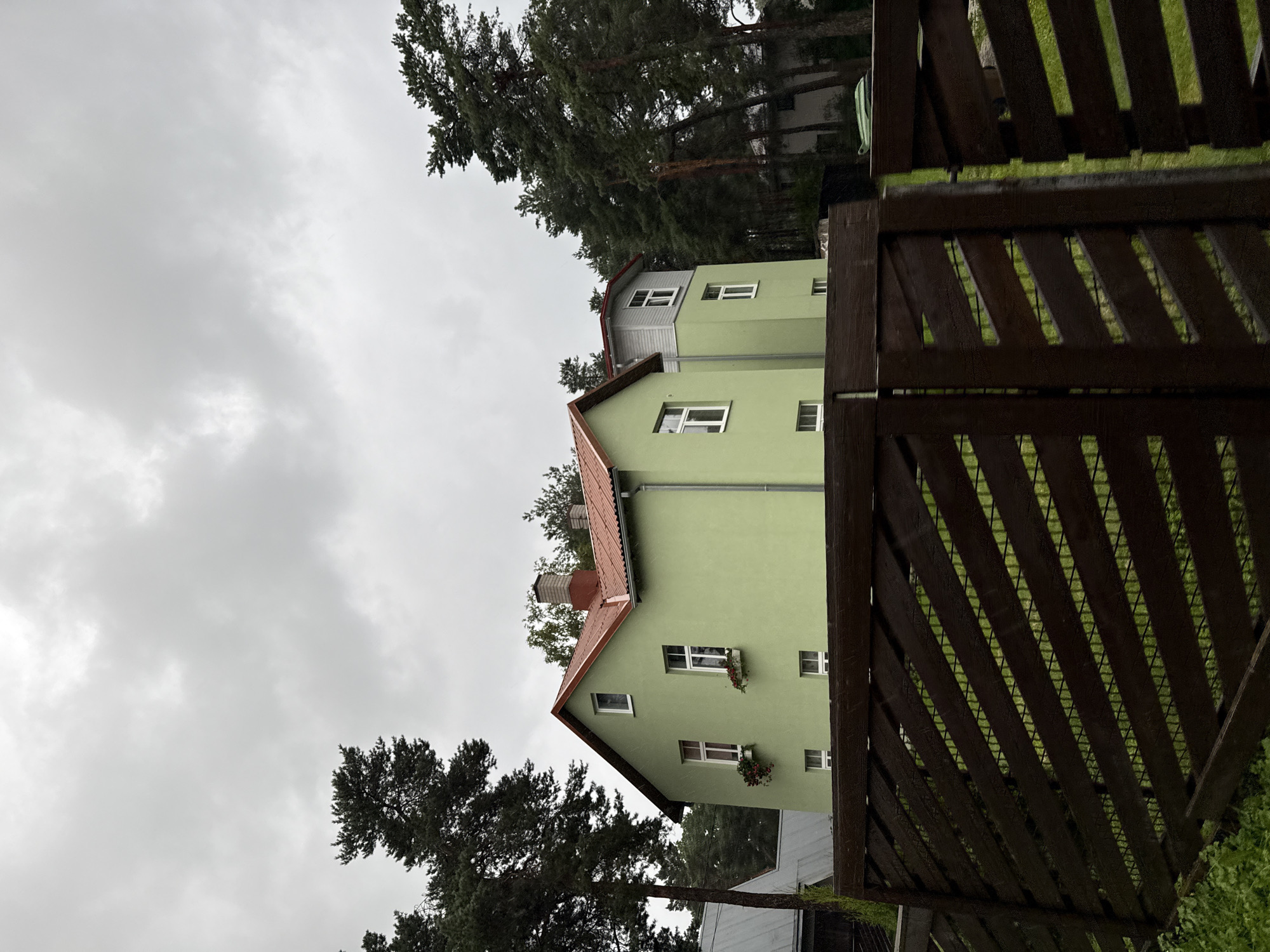 The image depicts a two-story light green house with a red tiled roof, situated in a densely wooded area. The house has multiple white-framed windows, some of which have small window boxes with red flowers. To the right of the main house, a portion of a slightly taller, gray-and-green structure is visible, also with white-framed windows and a red roof. A dark brown wooden fence with diagonal slats runs across the foreground, partially obscuring the view of the houses and the green lawn behind it. Tall pine trees surround the property, and the sky is overcast with thick gray clouds. The scene suggests a residential property in a natural setting, likely in a suburban or rural part of Tallinn, Estonia. No people are visible in the image.