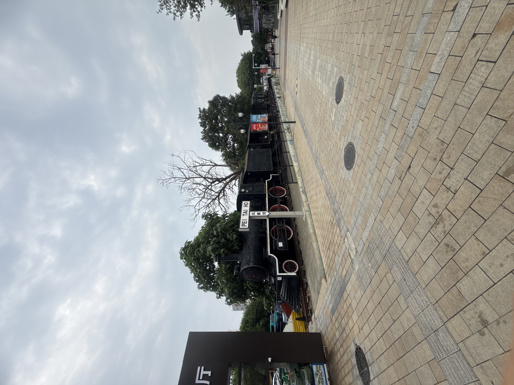The image shows a black steam locomotive exhibit on train tracks in an outdoor plaza. A signpost next to the locomotive reads "PEI TAI HO" in English and Chinese characters. The plaza has a patterned brick walkway with tactile paving for the visually impaired. Several trees are visible behind the train, and buildings are in the background. The sky is overcast. This scene is located at the Pei-tai-ho Railway Station in Qinhuangdao, China. There are no people prominently featured in the foreground, but a few individuals are visible in the distance walking along the plaza. The area appears to be a public space, possibly a historical exhibit or a recreated station.