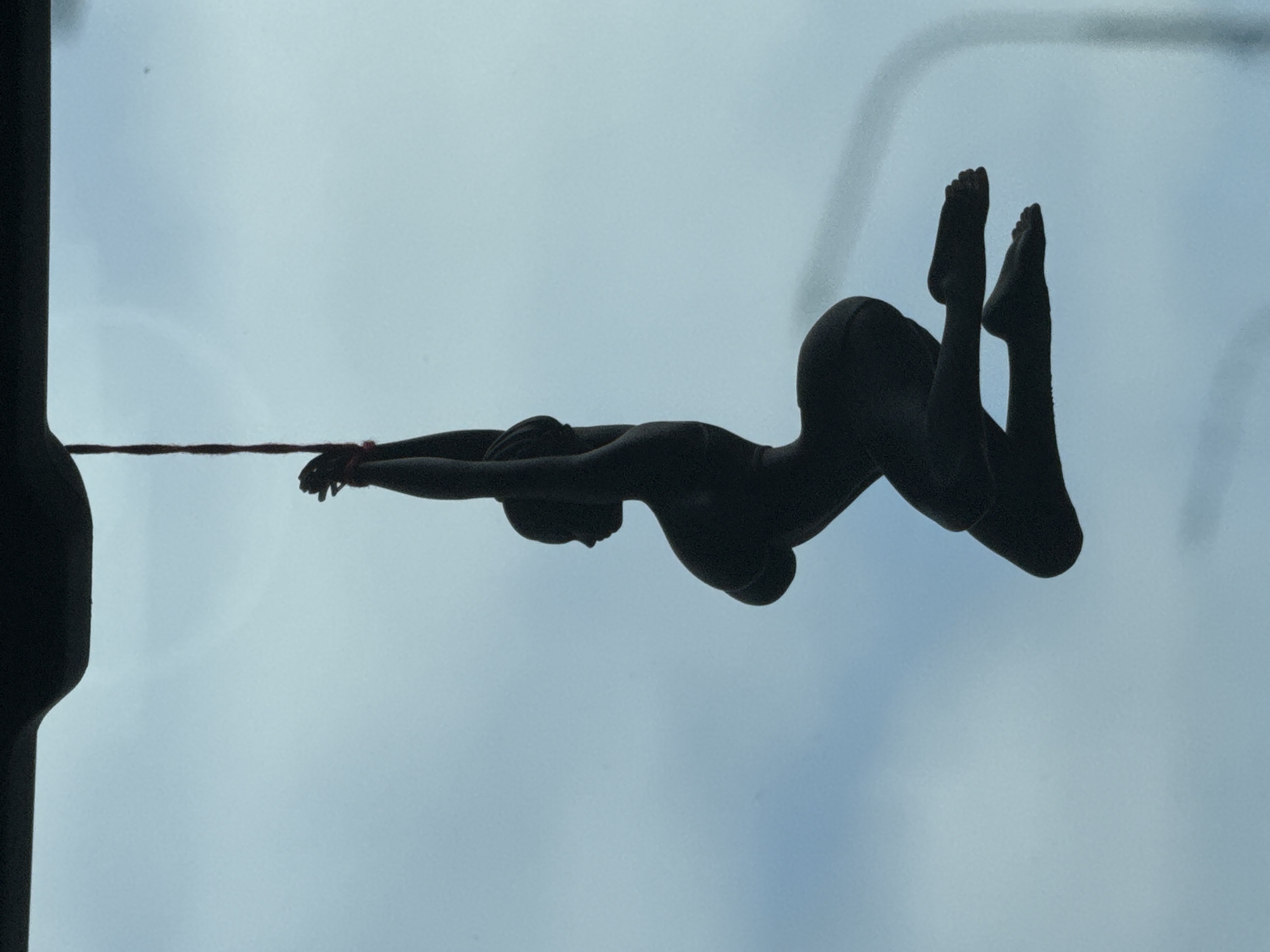 A silhouette of a nude female figure is suspended in the air against a pale blue sky. The figure is dark, obscuring any details of skin tone or features. It is depicted in a contorted pose, with arms raised and bound behind the head, and legs bent at the knees. The figure appears to be made of a solid material, likely plastic or resin, and is suspended by a thin red string tied around the wrists. The string is attached to an unseen object above, which casts a dark shadow across the top of the image. The sky is overcast, with diffused light and a faint vertical line on the right side of the frame, possibly a building or telephone pole. There are no discernible landmarks or environmental cues to suggest a specific location in Didino, Russia. The image is a close-up shot, focusing solely on the suspended figure and the sky.