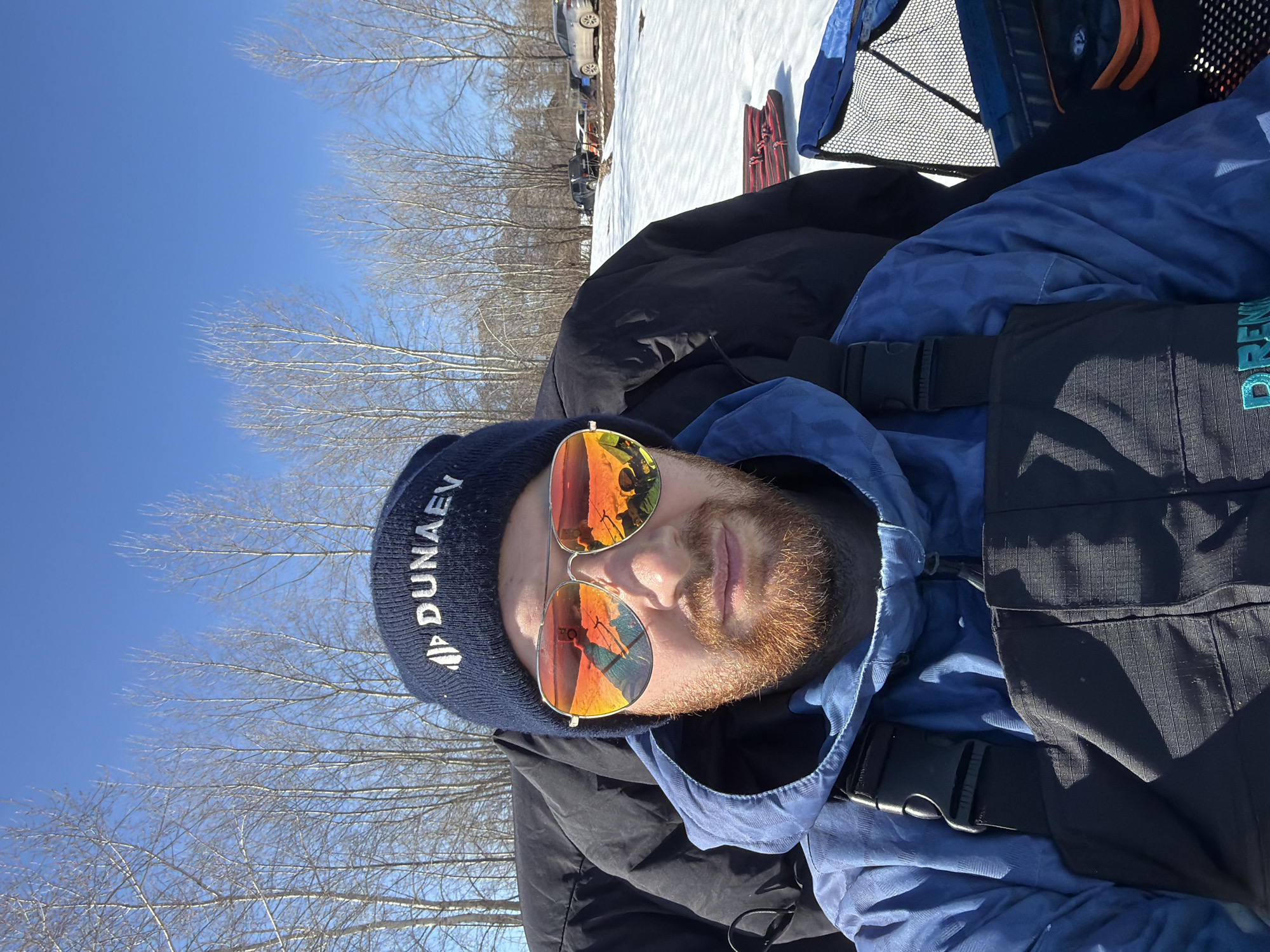 A selfie shows a person with a reddish beard and wearing sunglasses, a navy blue beanie with white lettering (partially obscured), a light blue hooded sweatshirt, and a dark jacket. The lenses of the sunglasses reflect what appears to be a snowy environment with people. A black backpack is partially visible. Bare trees and a bright blue sky are in the background. In the upper left corner, snowy ground with vehicles is visible. The hat reads "DUNAEV," a Russian surname. Given the name on the hat, it is possible the photo was taken in the city of Chaikovsky, Russia.