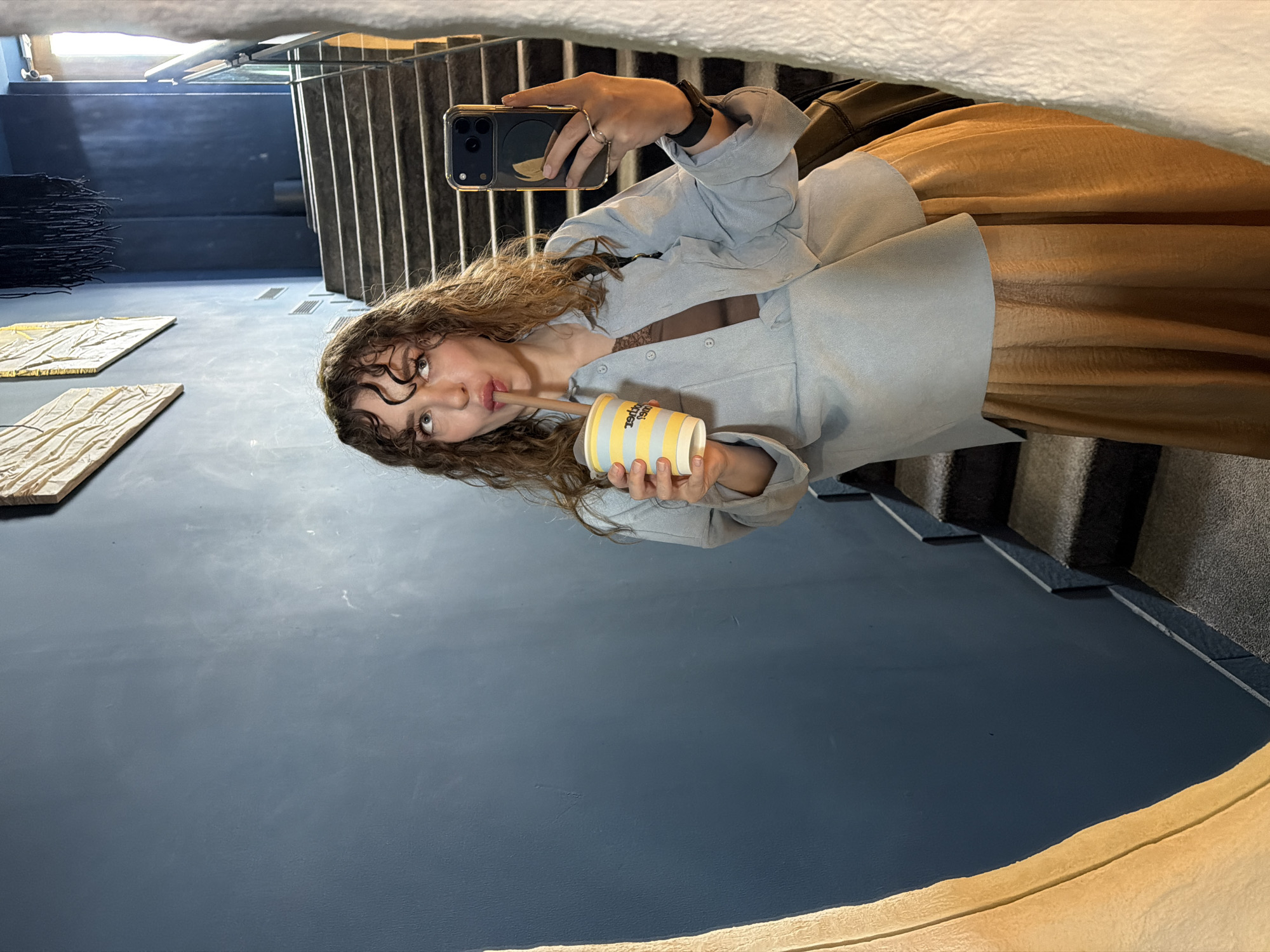 A young woman captures a selfie reflected in a mirror or highly polished surface within a contemporary indoor setting. She has long, curly, light brown hair and is seen from the chest up. She wears a light blue oversized button-up shirt over a darker, possibly lace, top, and a flowing brown/tan skirt or dress. On her left hand, she sports a ring and a watch, while holding a gold-colored iPhone with a clear case and a circular black accessory that appears to have a lemon design. Her right hand holds a yellow and white striped cup with a brown straw, from which she is sipping. The text "Życie Smakuje Lepiej" (Life Tastes Better) is visible on the cup.
The background shows a modern interior space, possibly a gallery or museum in Moscow, Russia. To the viewer's left, there are two textured, rectangular art panels on the floor and a dark, abstract, spiky art installation further back. To the viewer's right, a dark, angular staircase with metal railings ascends. The floor and walls are a uniform dark blue/grey, contributing to a sleek and minimalist aesthetic. The lighting is soft and even, typical of indoor environments. The woman's expression is neutral and focused on sipping her drink while taking the photo.