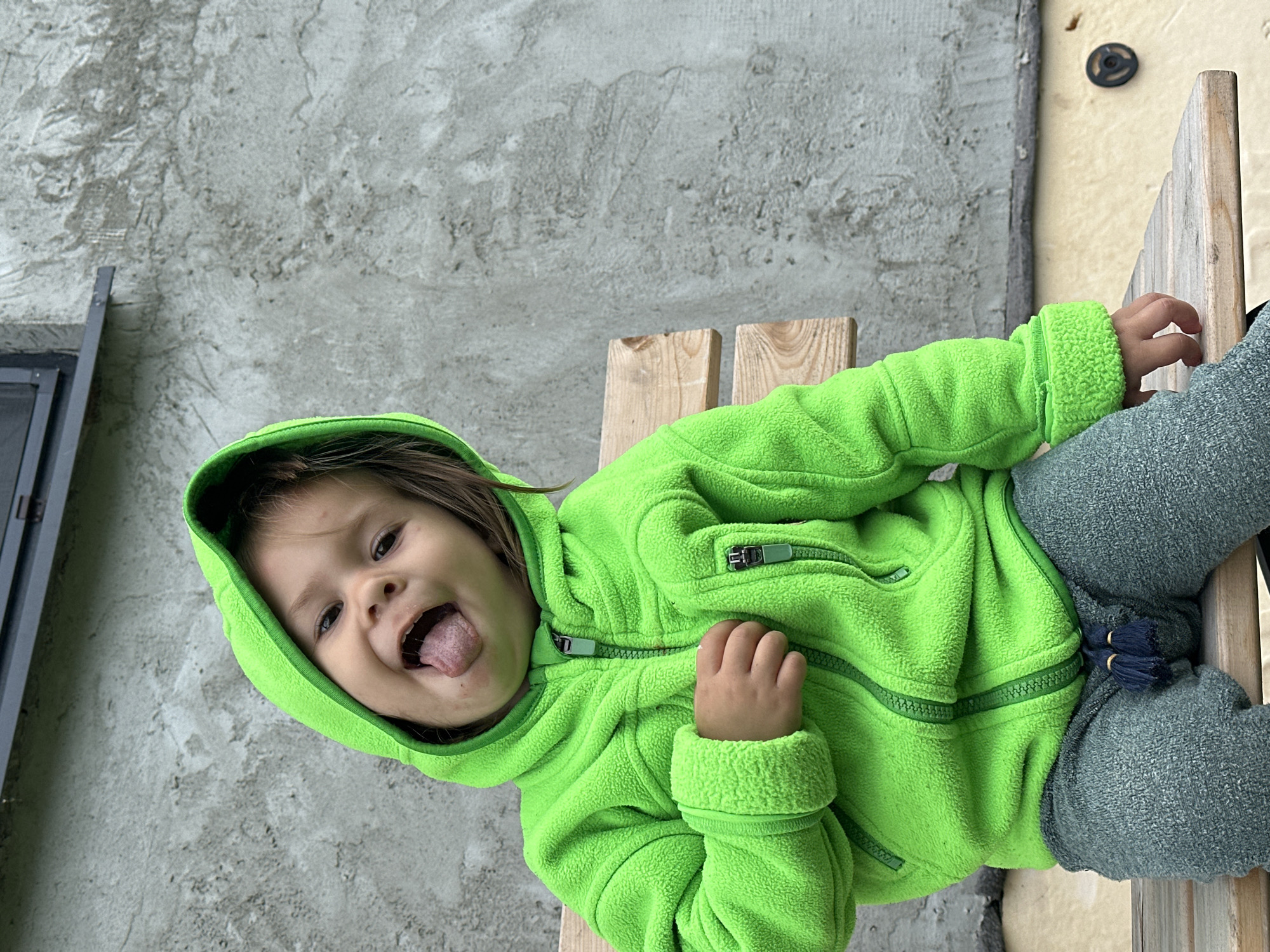 A child wearing a bright green hooded jacket and grey sweatpants sits on a wooden bench. The child is sticking out their tongue and looking at the camera. The background is a rough, grey concrete wall with a dark horizontal band across the top. The scene appears to be outdoors, possibly in a backyard or alleyway. There is no specific indication of the city of Kharnavy, Russia within the image.