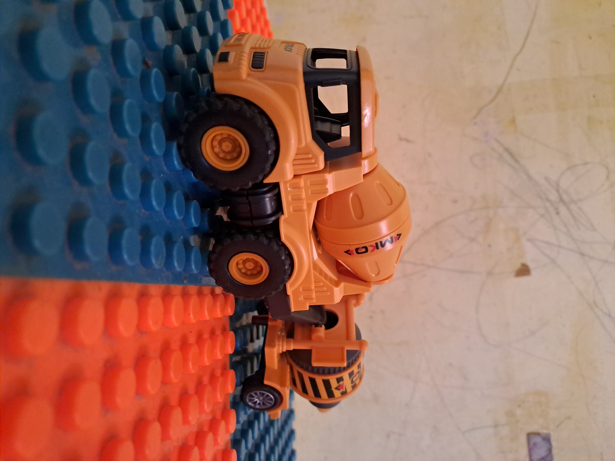 A close-up, eye-level shot depicts an orange toy cement mixer truck resting on a surface made of blue and orange plastic building blocks. The background is a light-colored wall with faint scribbled markings. The truck is positioned diagonally across the frame, with its front end on the left and its rear extending towards the right. The truck's main drum is tilted, and the cab is visible with its windows. The building blocks are arranged in a grid pattern. The lighting appears to be coming from the front and slightly to the left. There are no people visible in the image.