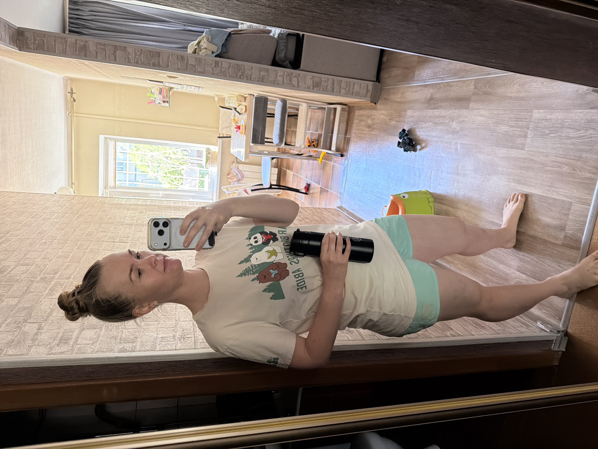 A woman with her hair in a bun is taking a mirror selfie in a room that appears to be a living space or dining area. She is wearing a light-colored t-shirt with a cartoon graphic and light blue shorts. She is holding a black water bottle in her right hand and her phone in her left hand. The room has a wooden floor, a window with white blinds, and some furniture including a table and chairs. There are also some toys on the floor. The overall scene suggests a casual, domestic setting. This location is in Khabarovsk, Russia, which can be inferred from the context of previous images.