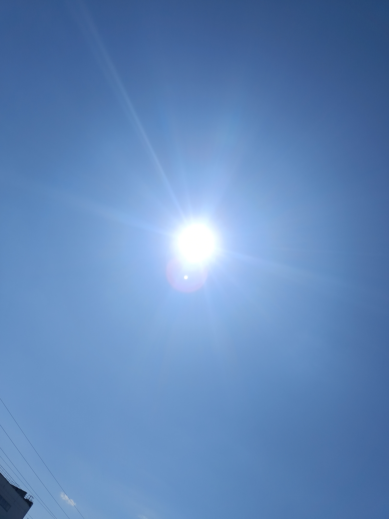 The image shows a clear, bright blue sky with the sun centered. The sun has a bright glare and light rays emanating outwards. In the lower left corner, there's a glimpse of a building and some power lines. I cannot determine the location of this image in the city of Chaikovsky, Russia.