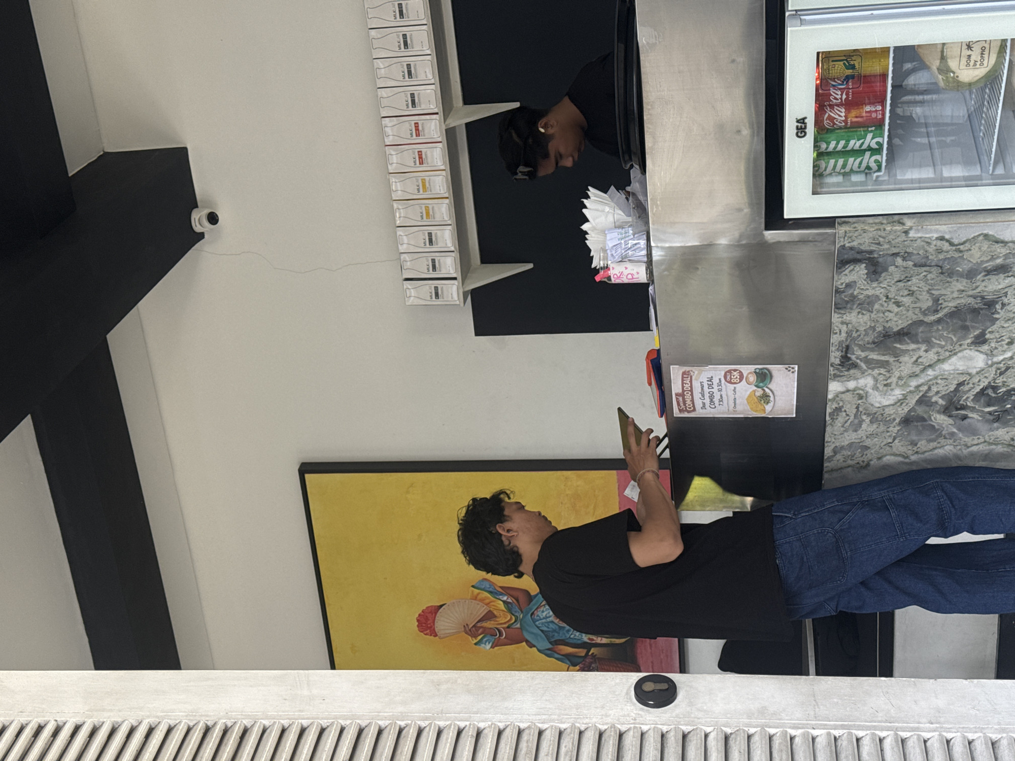 The image shows the interior of a shop. A man with curly black hair and wearing a black T-shirt and blue jeans is standing, holding a smartphone, appearing to interact with an employee behind a counter. The counter is equipped with a drinks refrigerator displaying brands like Sprite and Coca-Cola. The refrigerator is labeled with 'GEA'. Shelves displaying rows of what appear to be 'MILKLAB' products are behind the employee. A painting depicting a woman with a fan adorns the wall behind the customer. A security camera is mounted on the wall near a structural element. A sign advertising a 'Social Combo Deal' is visible on the counter. The location is North Kuta, Indonesia, as indicated by the 'COMBO DEAL' price being in 'K' currency.
