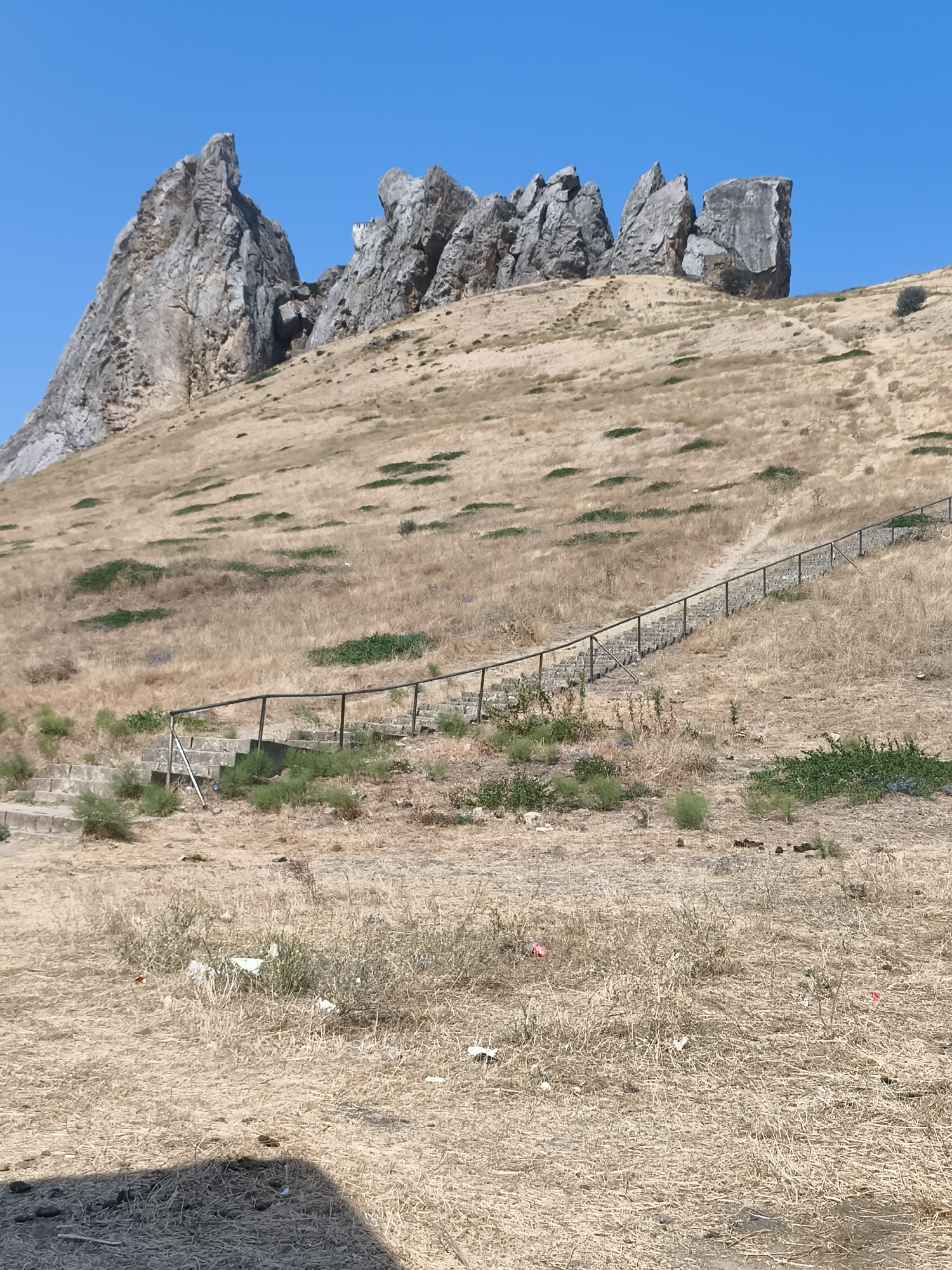 In Siazan, Azerbaijan, a rocky mountainous terrain under a clear blue sky is depicted. Dry, golden grass covers the sloping hills, with scattered patches of green vegetation. A stone staircase with a metal railing winds its way up the side of the hill. At the summit, jagged rock formations rise prominently. The foreground shows a dry, grassy area with some scattered litter. A shadow is cast on the lower left corner of the image.