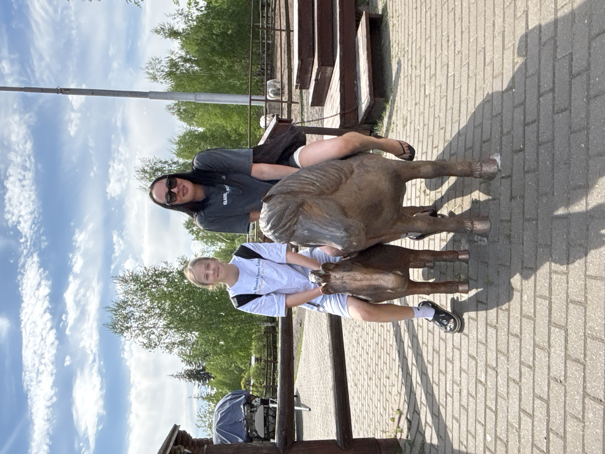 Two females are posing on a bronze horse sculpture on a brick-paved surface. The female on the top horse wears sunglasses and a gray t-shirt. The female on the lower horse wears a white t-shirt with black accents. In the background are trees, sky with clouds, a wooden fence, and stairs. This is likely at the Izhevsk Zoo, Russia.