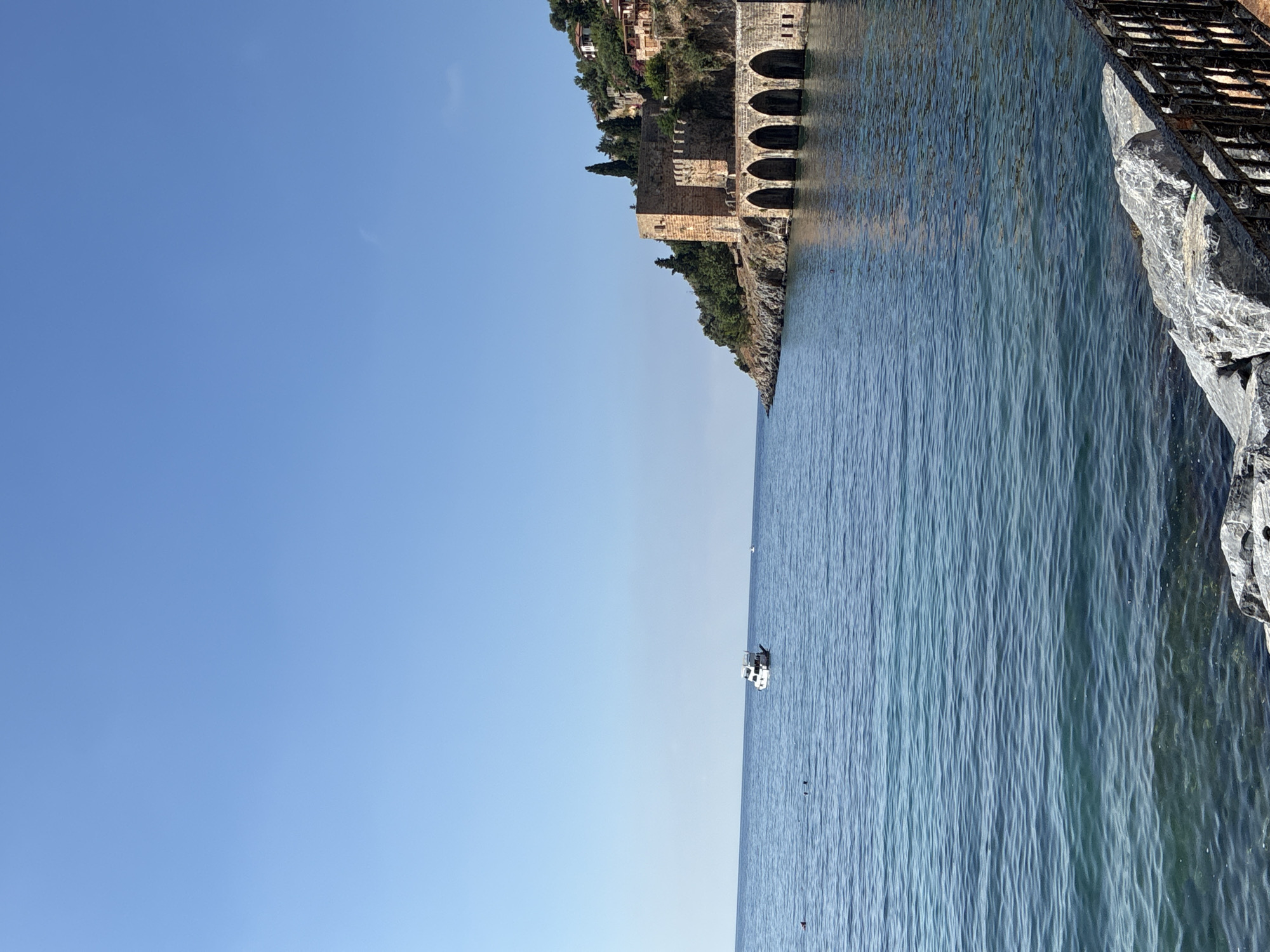 The image depicts a coastal scene in Alanya, Turkey. In the foreground, the clear, blue water of the Mediterranean Sea laps against a rocky shore. A black metal railing runs along the edge of the shore. In the distance, a white boat is visible on the water, and further out, two small figures appear to be swimming. On the right side of the frame, an ancient stone structure, likely part of the Alanya Red Tower or shipyards complex, is perched on a cliff overlooking the sea. This structure features arched openings at its base and is surrounded by lush green trees. The sky above is a clear, bright blue, indicating a sunny day.