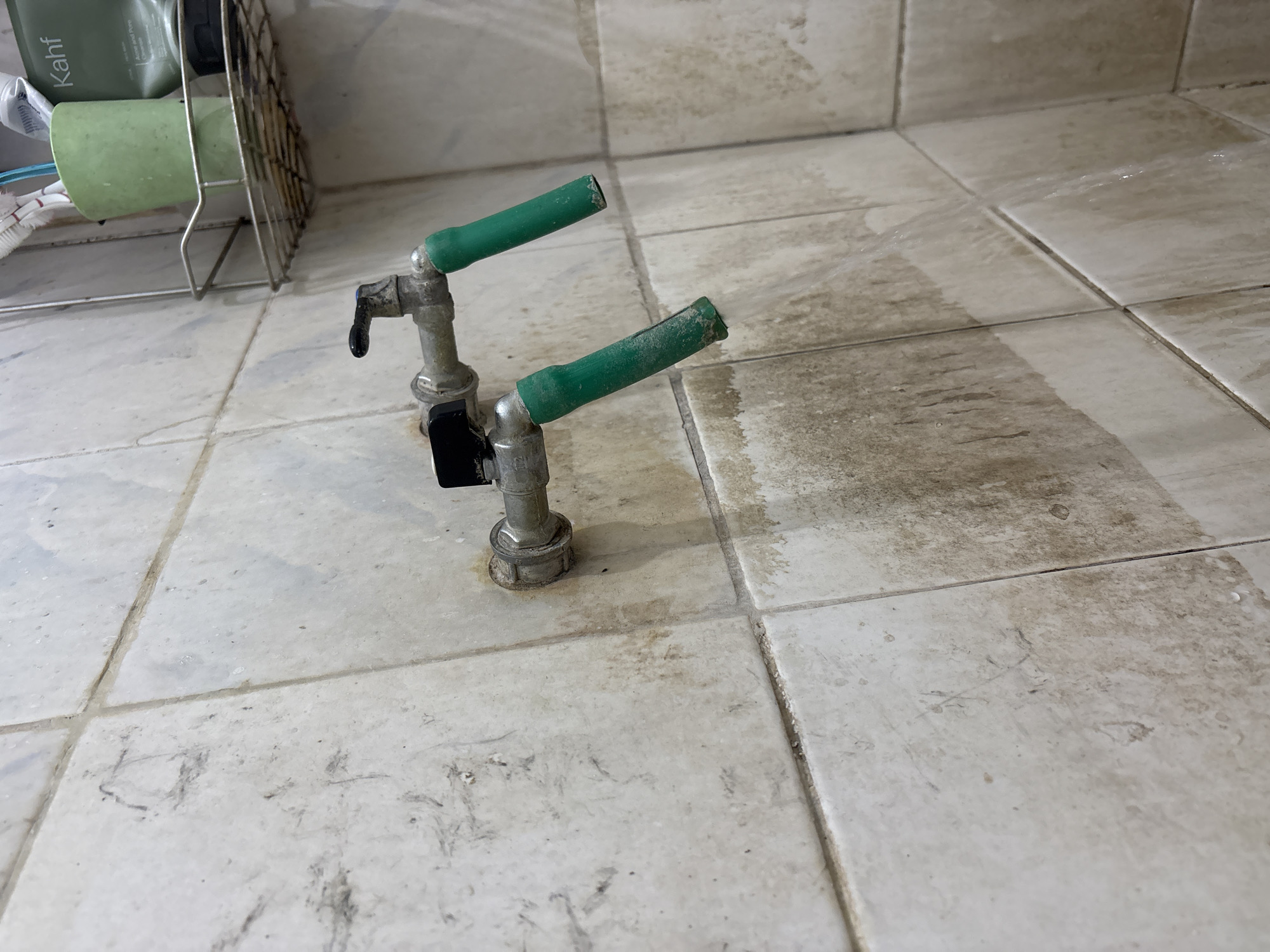 The image shows two metal faucets with green handles mounted on a tiled wall. The tiles are off-white and appear to be dirty, with dark streaks and water stains. The faucets are positioned horizontally, with the handles pointing downwards and slightly outwards. One faucet has a black valve control. A shelf with toiletries, including toothbrushes and tubes of product, is visible in the upper right corner. The environment suggests a bathroom or utility area. The specific location in Palimanan, Indonesia, cannot be determined from the visual information alone.