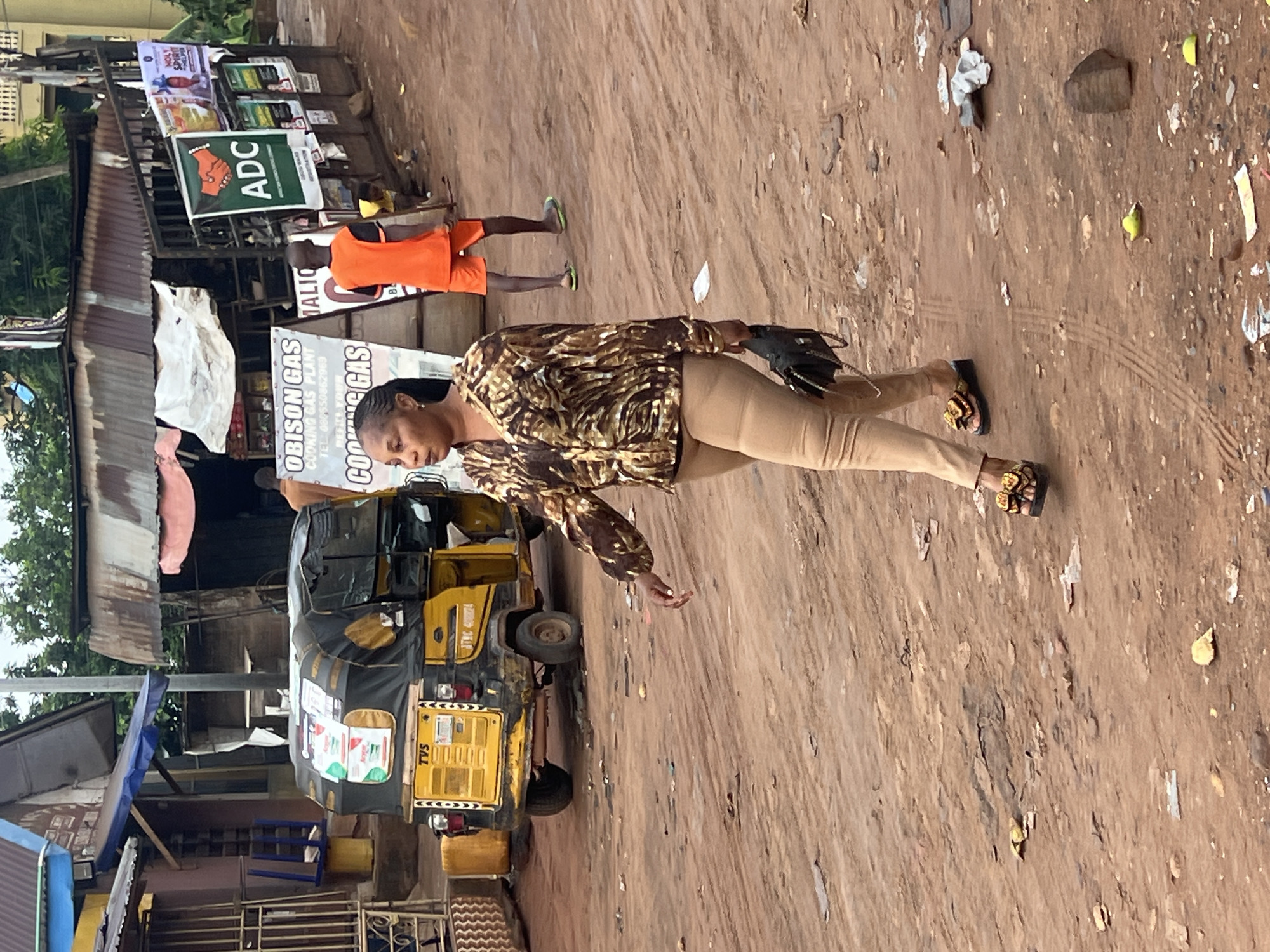 The image shows a woman walking on a dirt road in Onitsha, Nigeria. She is wearing a patterned blouse, khaki pants, and sandals, and carrying a dark object. A yellow auto rickshaw (TVS branded) with a black canopy sits parked to her left, near a shop advertising cooking gas ("OBISON GAS" and "COOKING GAS PLANT"). A man wearing orange shorts is also seen in the background, walking near a sign with the letters "ADC". The surrounding environment includes buildings with corrugated iron roofs and various posters and signs attached to structures. The ground is covered in dirt and scattered debris.