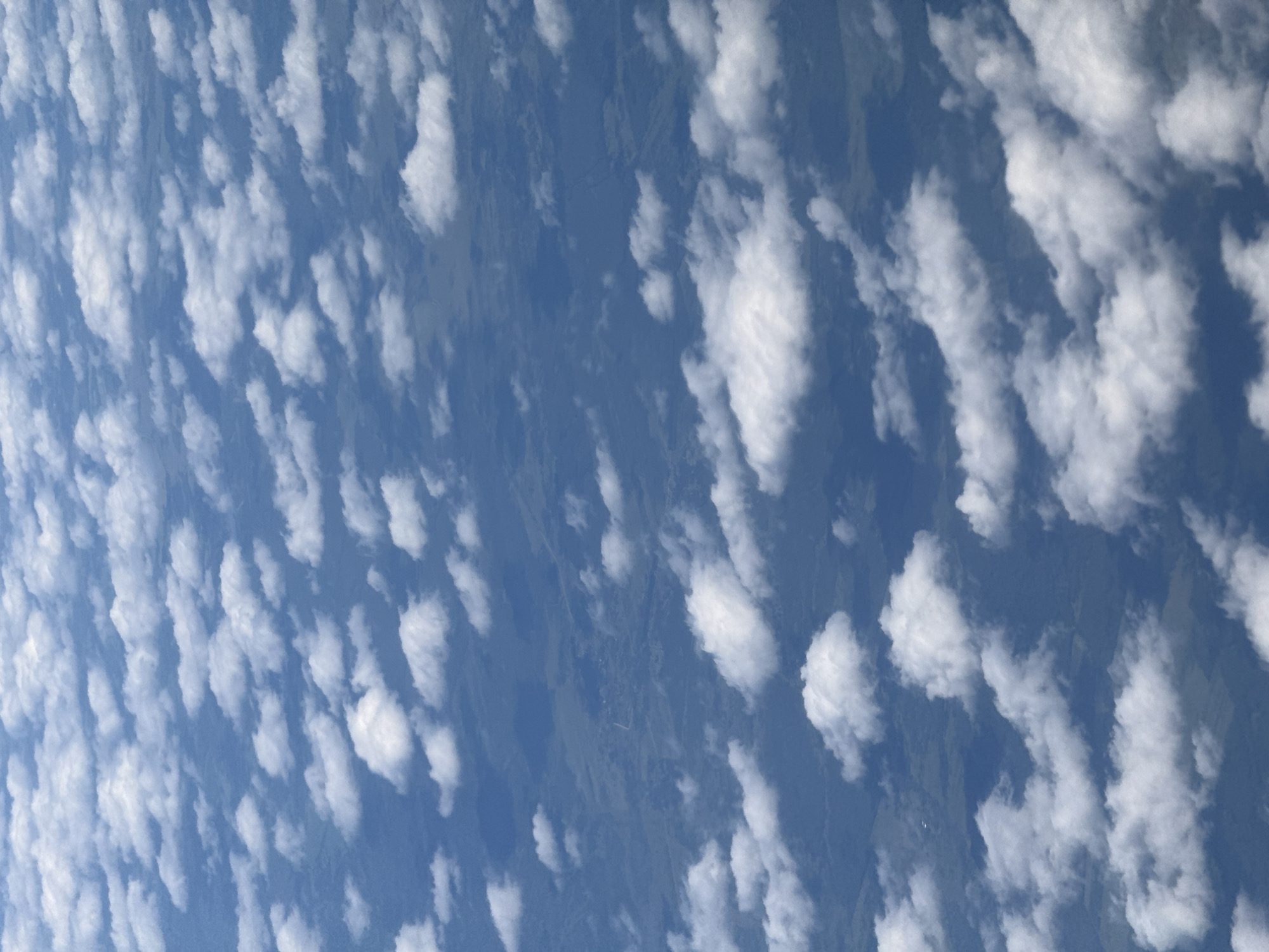 This is an aerial photograph looking down on scattered cumulus clouds and the landscape below. The ground appears to be rural, with patches of green fields and possibly some trees. There are no discernible buildings or man-made structures visible. The photograph was taken from a high altitude, likely from an airplane, as indicated by the perspective and the presence of clouds. The specific location in Kazan, Russia, cannot be determined from the visual information alone.