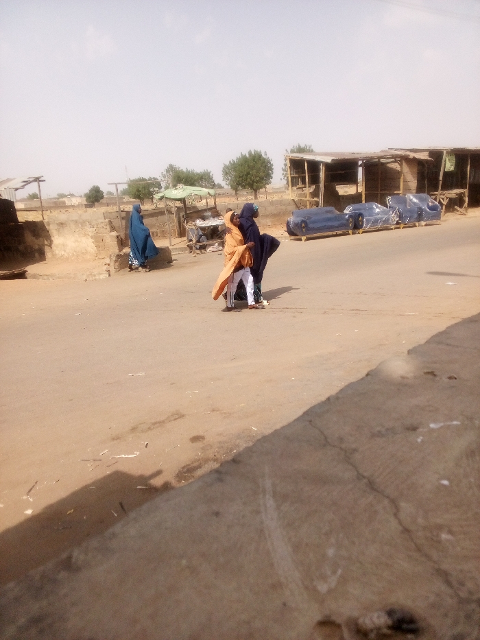 This image captures a street scene in Tudun Murtala, Nigeria, under bright, possibly midday, lighting conditions. The foreground consists of an unpaved road or dirt-covered asphalt surface, with a low concrete barrier or curb on the right.
Three individuals are visible. On the left side of the road, a person wearing a full-length blue garment stands facing left, positioned near a dirt mound and a rudimentary structure. In the center-right of the road, two individuals are in close proximity, appearing to walk or stand together. One person wears an orange or light brown full-length garment over white trousers, while the other wears dark blue or black full-length attire over what appears to be green trousers.
The environment includes several structures. On the immediate right, a series of open-fronted, wooden stalls with corrugated metal roofs line the road. In front of these stalls, several pieces of blue upholstered furniture, possibly sofas or chairs, are visible, individually wrapped in clear plastic and resting on wooden pallets. Further in the background, more distant buildings and scattered green trees are visible under a hazy sky. Debris, including small pieces of litter and dark stains, is present on the road surface.
