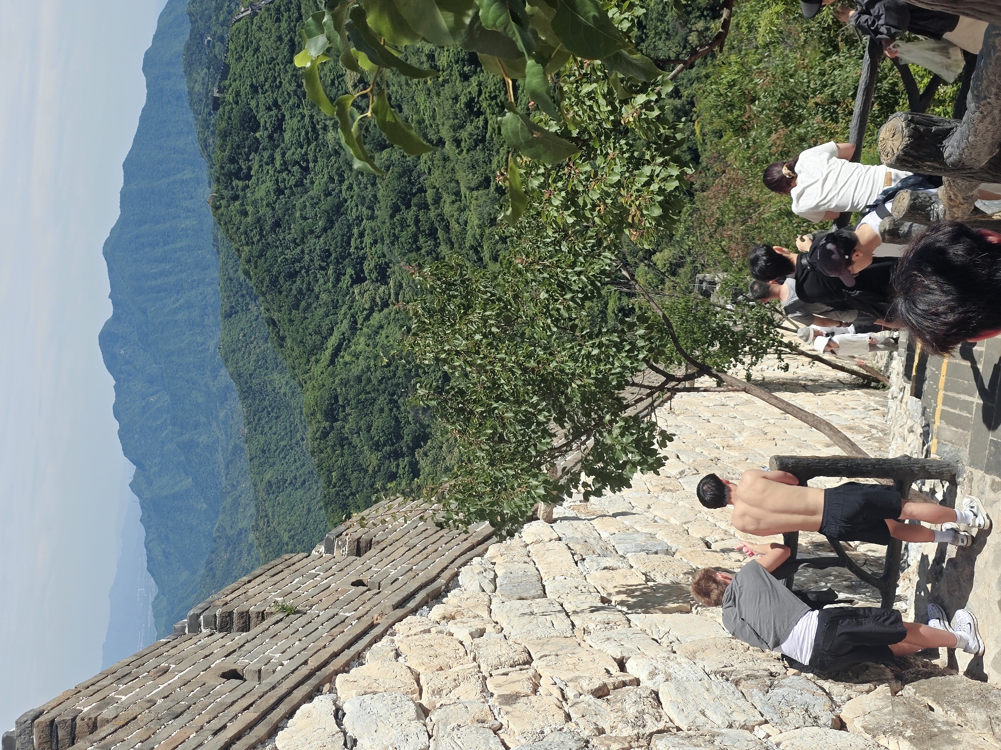 This image depicts a group of people at the Great Wall of China in Huairou District, China. The scene is set on a bright, sunny day with clear skies, suggesting it's mid-morning or afternoon. The Great Wall stretches across a mountainous, densely forested landscape, with layers of hazy blue mountains receding into the distance. The wall itself is constructed from weathered stone and brick, with a wooden railing in the foreground.
In the foreground, two young men are engaged in physical activity. One, shirtless, is performing push-ups with his hands on the wooden railing, while the other, wearing a grey shirt and black shorts, is in a plank position on the stone ground. Both are wearing athletic shoes.
Further back, several other individuals are visible, some looking out at the scenic vista, others walking along the wall. One person in a white shirt is leaning on the railing, observing the view. The people appear to be tourists, enjoying the historical site and its natural beauty. The overall atmosphere is one of leisure and exploration. No visible text is present in the image.