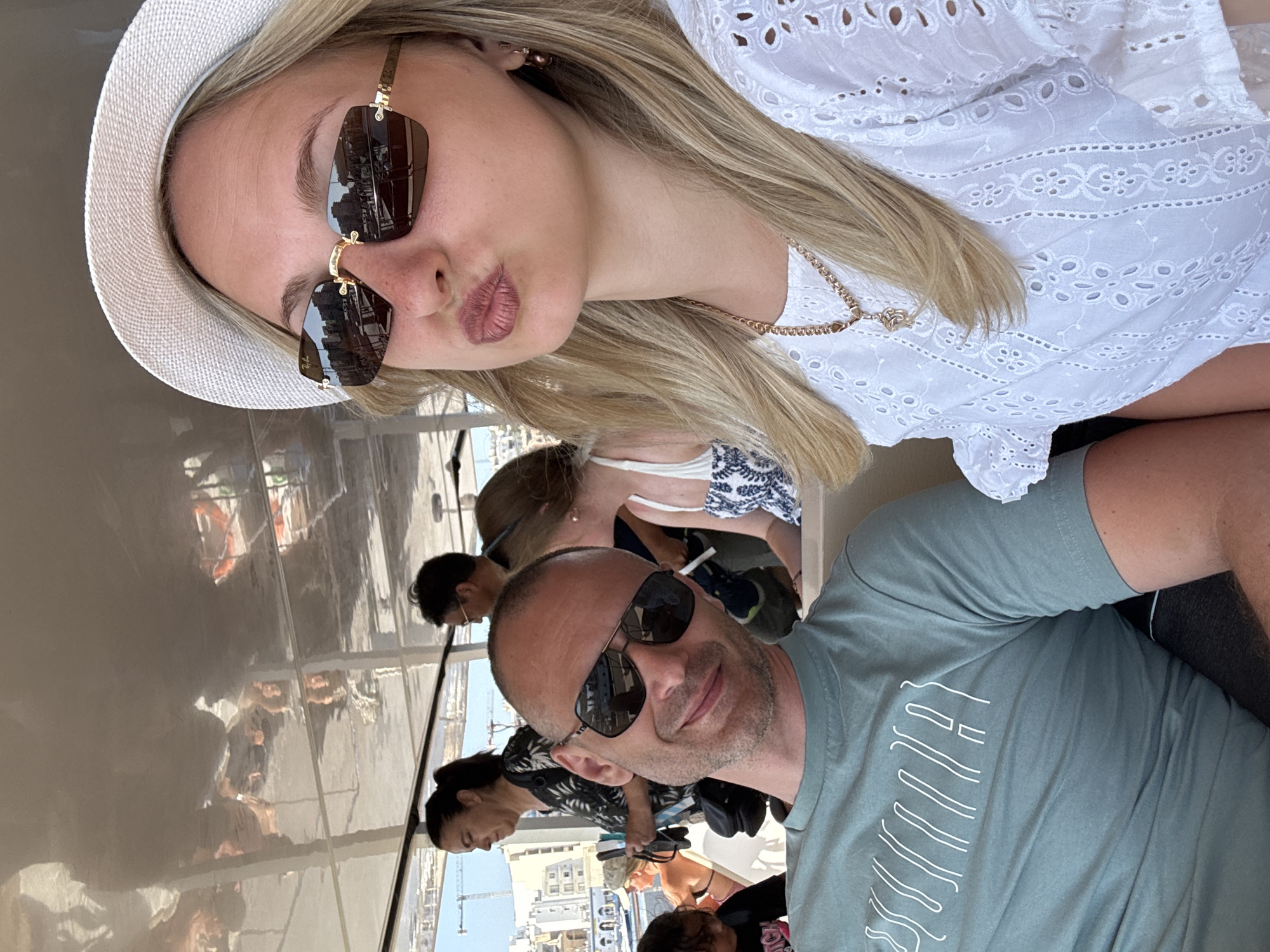 The image shows a selfie taken on a boat tour, likely in a European city given the architecture reflected in the woman's sunglasses. In the foreground, a woman with blonde hair wearing a white hat and a white embroidered top is posing for the camera with her lips puckered. She is wearing sunglasses and a delicate necklace. To her left, a man with a shaved head wearing sunglasses and a teal t-shirt is looking at the camera with a smile. In the background, other passengers are visible, as well as reflections of buildings on the boat's ceiling. The overall scene depicts a casual outing on a sunny day.