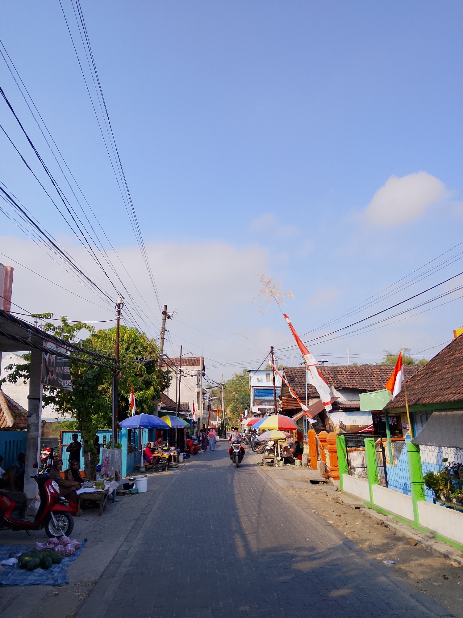 The image depicts a street scene in Batokan, Indonesia, on a clear, sunny day. The street is paved with interlocking bricks, and on both sides, there are buildings with tile roofs, some of which appear to be shops or homes. Several Indonesian flags, predominantly red and white, are displayed, suggesting a national celebration or patriotic display.
Along the street, several vendors have set up stalls under colorful umbrellas, selling goods. In the foreground on the left, a red scooter is parked next to a stall displaying watermelons and onions. People are seen sitting, standing, and interacting at these stalls. Further down the street, a motorcycle is being ridden, and other people are walking.
The sky is a bright blue with a few scattered white clouds. Overhead, a dense network of electrical wires crisscrosses the sky, supported by utility poles. The overall atmosphere appears to be one of a bustling local market or a residential street with commercial activity.