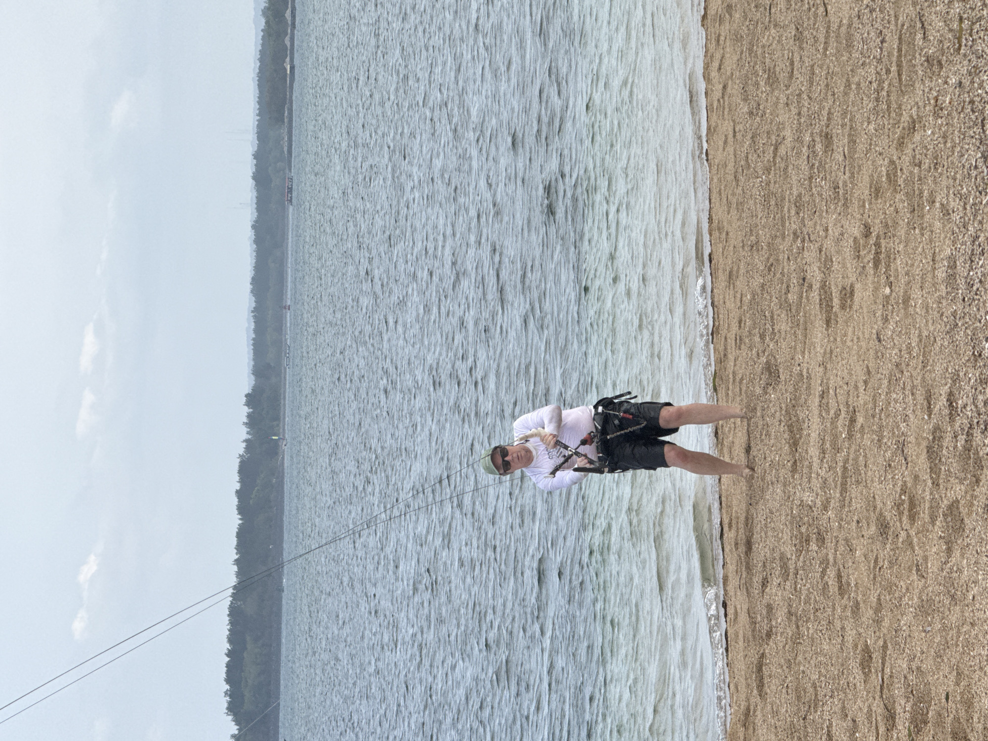 A person stands on a sandy beach, facing the camera and holding onto kiteboarding equipment. The person is wearing a white long-sleeved shirt, black shorts, a green cap, and sunglasses. They are wearing a kiteboarding harness around their waist and holding a control bar with both hands. The person's feet are bare and appear to be in shallow water at the edge of the beach. The beach is covered in light brown sand with some small shells. The ocean behind the person is choppy and a light blue-gray color. In the background, across the water, is a distant treeline and a hazy sky. The location is the shore of Denpasar, Indonesia.