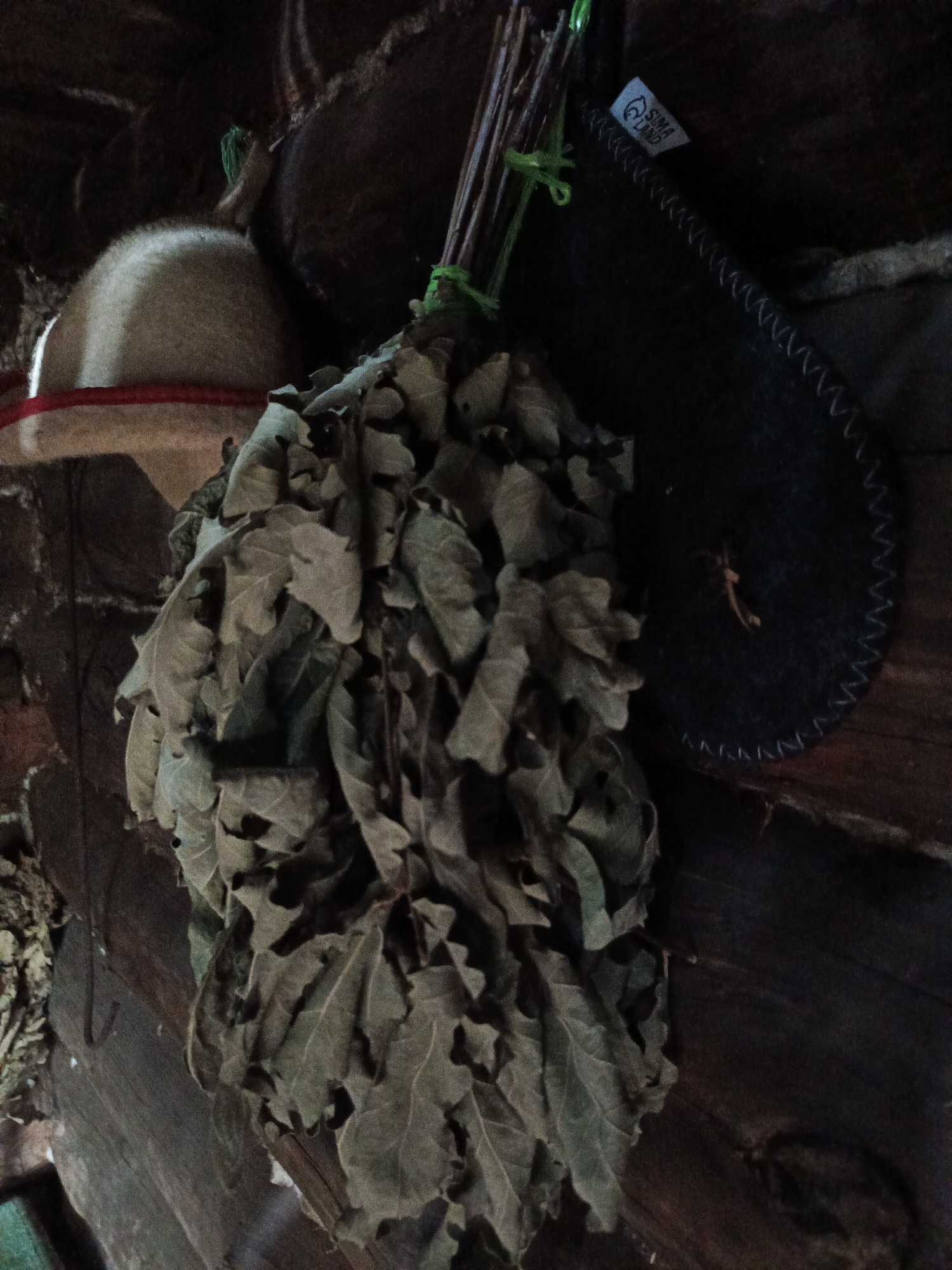 A dried leafy bath whisk hangs against a wooden wall. The whisk, made of oak leaves tied with green string, is on the left side of the image. To the right of the whisk is a dark, round object with white stitching along its edge. A small white tag with the logo "SIMA LAND" is attached to the object. A portion of a beige, furry object with a red rim is visible on the upper left. The background consists of dark, rough-hewn logs. The scene appears to be indoors.