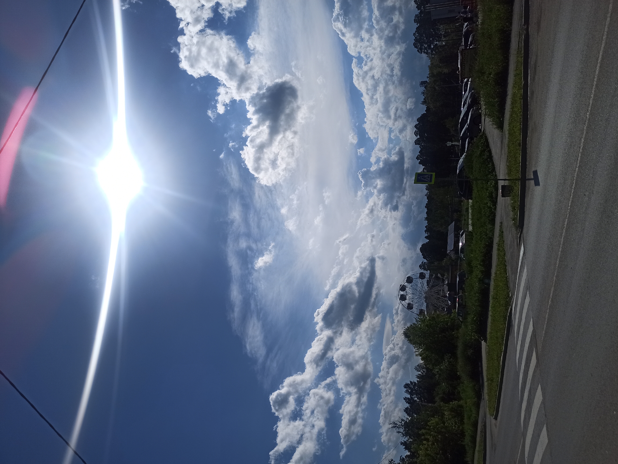 The image captures a sunny day in Asbest, Russia, with a bright sun shining in a blue sky dotted with fluffy white clouds. A power line runs diagonally across the upper left corner of the frame, with a lens flare from the sun extending downwards. In the background, a Ferris wheel is visible, hinting at a possible amusement park or recreational area. To the right of the Ferris wheel, a line of parked cars is seen next to a grassy area and trees. A road with a pedestrian crossing is in the foreground, suggesting an urban or suburban setting. The overall impression is one of a bright and clear day, possibly during the daytime, with recreational elements and everyday infrastructure present. No people are clearly discernible, but their presence is implied by the cars and the pedestrian crossing. The weather appears pleasant and sunny.