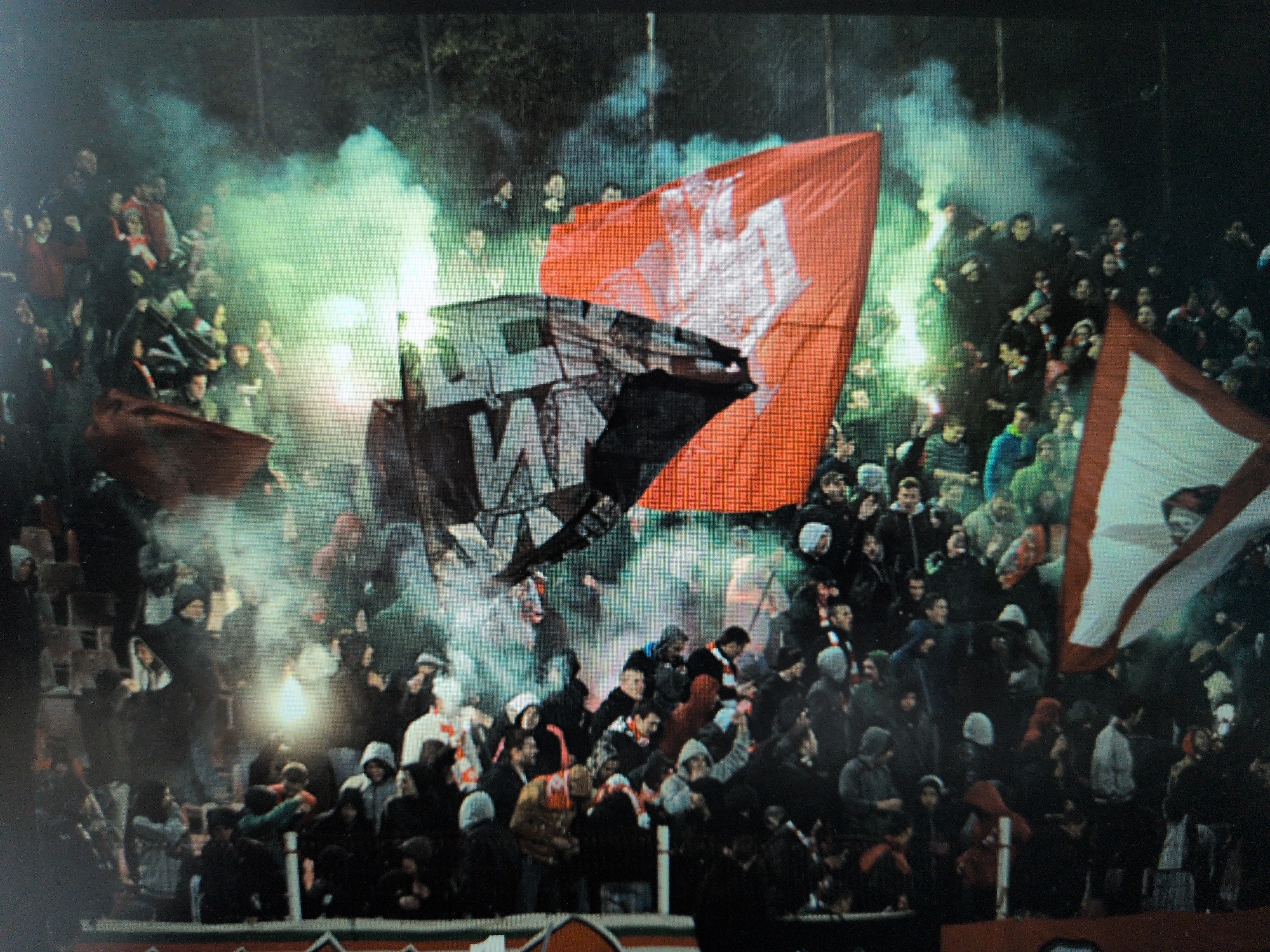 A vibrant and intense scene unfolds in what appears to be a football stadium or arena, filled with a large crowd of spectators. The image captures a moment of fervent support, characterized by the use of flares and large flags.
The primary subjects are numerous people, mostly men, densely packed together in a spectator stand. They are dressed in casual clothing, including hoodies, jackets, and hats, suggesting cool weather. Several flares are active within the crowd, emitting bright light and thick green smoke that permeates the air, creating a dramatic, hazy atmosphere.
Dominating the center of the frame are two large flags. One is a bright red flag, partially unfurled, featuring a white symbol or emblem that is difficult to discern clearly, possibly a stylized figure. Behind and overlapping it is a black flag with bold white block letters. The visible text on the black flag appears to begin with "U" followed by a large, blocky letter that resembles an "N" or "M". Other letters are partially obscured by folds and smoke. On the far right, part of a white flag with red stripes is visible.
The event is likely a sporting match, given the stadium setting and the passionate display of fan culture, commonly associated with "ultras" or dedicated supporter groups. The time of day appears to be night or late evening, as the flares cast significant light and the surroundings are dark, adding to the dramatic effect. The overall mood is one of excitement, energy, and collective fervor. No specific location cues are visible beyond the general stadium structure.