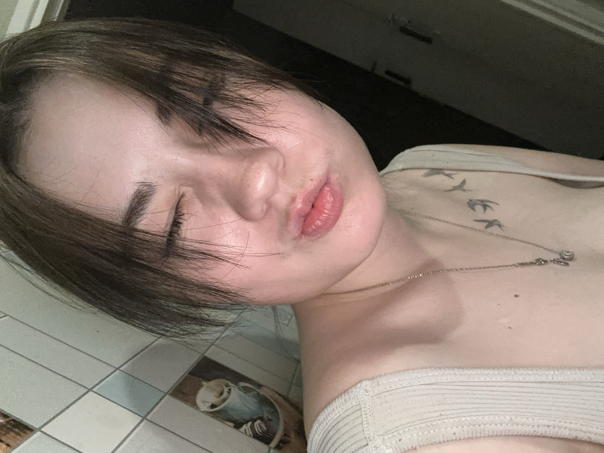 The image shows a close-up, selfie-style shot of a young person with their eyes closed and lips puckered, as if blowing a kiss. They have short, dark brown hair that falls across their forehead. They are wearing a light-colored, ribbed tank top and a necklace with a pendant. A tattoo of what appear to be birds in flight is visible on their upper chest. The background appears to be a tiled wall, possibly in a bathroom or kitchen, with a decorative tile featuring a mug of coffee. The person is positioned in what could be a residential setting. The metadata does not contain information about the specific location in Chaikovsky, Russia.