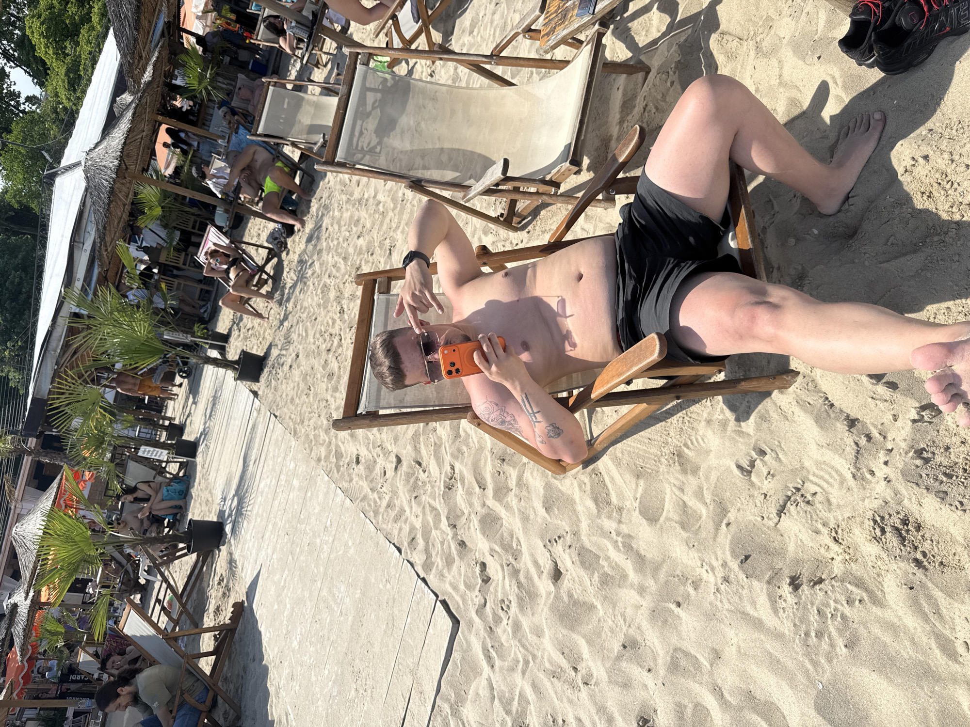 The image depicts a vibrant outdoor scene at a beach establishment in Varna, Bulgaria. The foreground is dominated by a male individual seated in a wooden beach chair on light-colored sand, facing slightly away from the camera. He is shirtless, wearing black shorts and sunglasses, holding an orange-cased smartphone in his left hand, and making a hand gesture with his right hand (index and pinky fingers extended). Tattoos are visible on his left bicep. His bare feet are partially submerged in the sand, with a pair of black and red sneakers resting on the sand to his right.
Behind the foreground subject, a wooden boardwalk extends diagonally across the sandy beach. Potted palm trees are strategically placed along the boardwalk and in the sand. Numerous wooden beach chairs, some occupied and some empty, are scattered throughout the midground. Several thatched umbrellas provide shade over groups of people relaxing in chairs or seated at small tables. A man with long dark hair and a beard is visible on the left, seated in a chair near the boardwalk, appearing to hold a cigarette. Further right, a woman in a bikini and sunglasses lies in a beach chair under an umbrella, while another man in yellow swim trunks sits in a nearby chair.
In the background, a white-roofed building, likely a bar or restaurant, stands behind the umbrellas. A sign with the word "BACARDI" is partially visible on the building's facade. Additional people are present within and around the building and shaded areas. The far background shows dense green foliage under a bright sky, suggesting a natural tree line beyond the developed beach area. The scene is brightly lit by natural sunlight, casting distinct shadows.