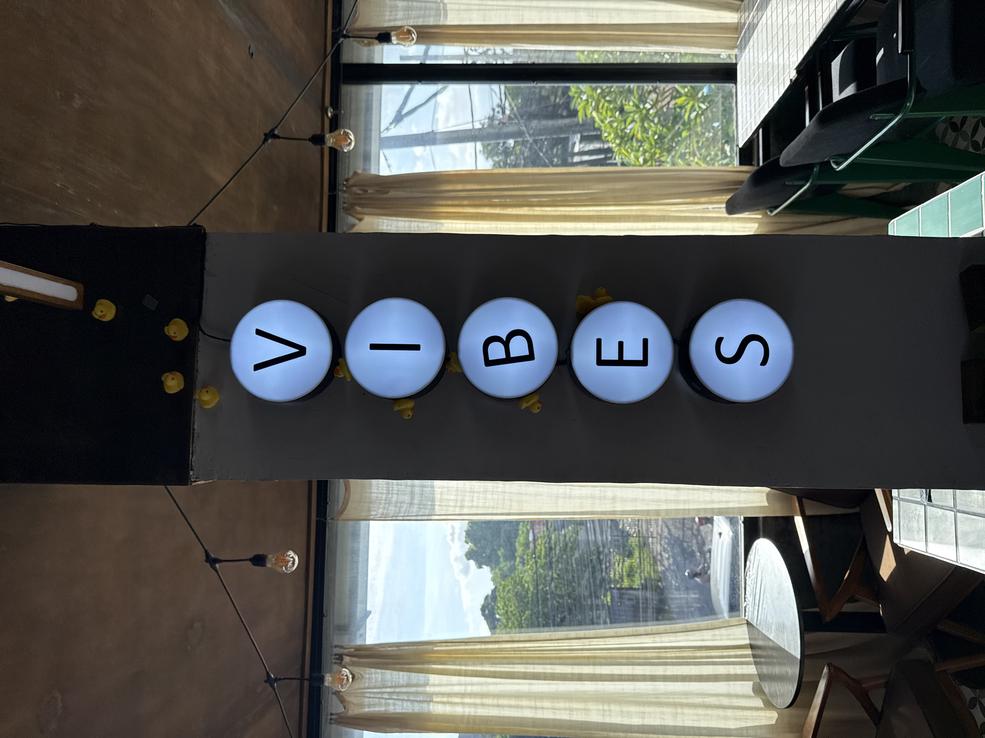 Captured from an unconventional angle, the image presents a room scene, likely from a commercial establishment, situated in Uluwatu, Indonesia. The focal point is a sign composed of five circular lights, each illuminating a letter to spell out "VIBES". Small yellow rubber ducks are visible atop the lights. The sign is affixed to a grey structure near windows. Through the windows, greenery and buildings typical of an urban environment are visible. Translucent curtains adorn the windows, softening the natural light. String lights with Edison-style bulbs stretch across the ceiling. The presence of furniture, including tables and chairs, suggests a cafe or similar establishment. The image is taken from a low angle, giving a unique perspective of the scene.