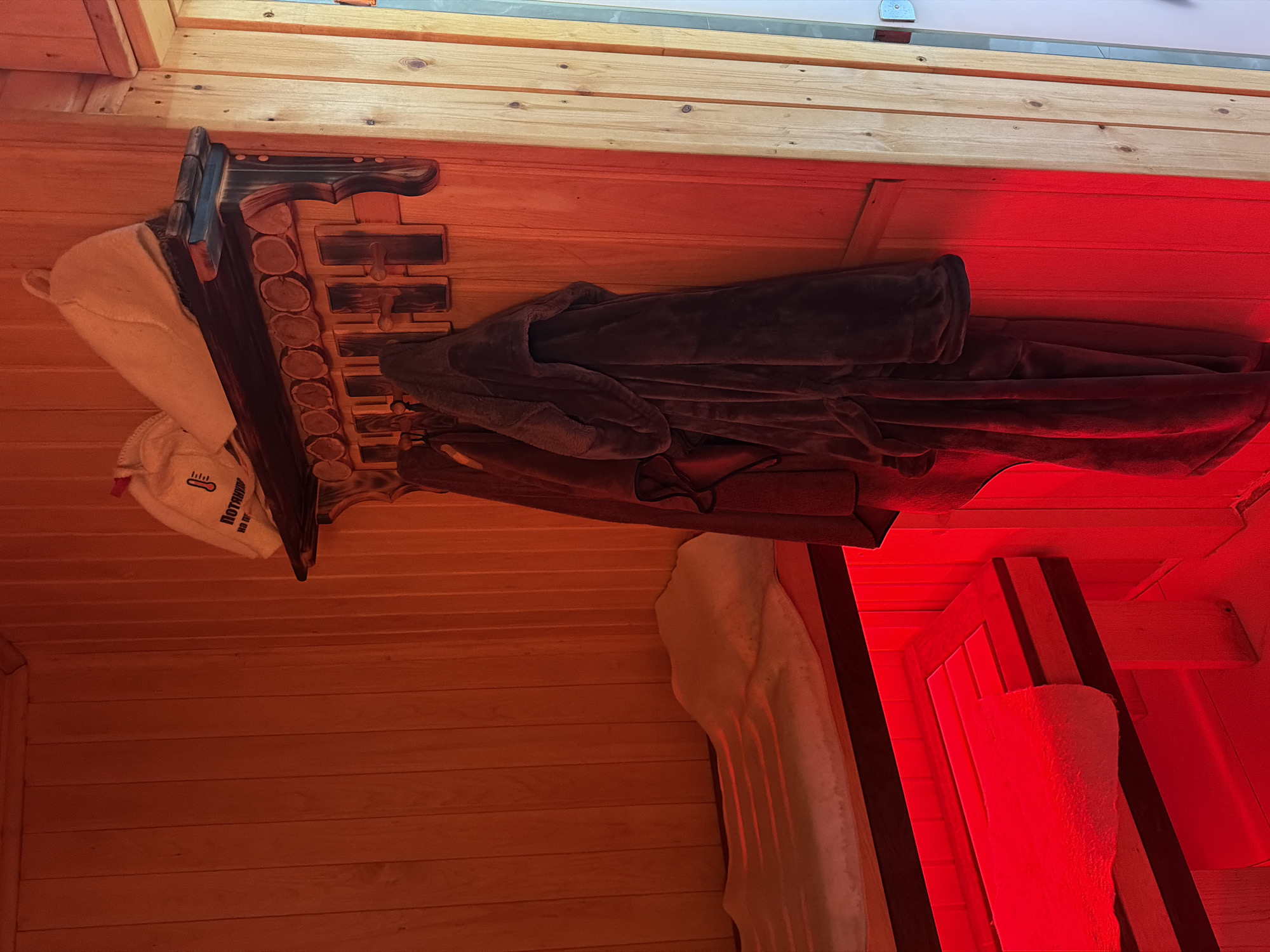 The image shows the interior of a sauna room, likely located in Orenburg, Russia, given the presence of what appears to be Russian text on a bag on the shelf. The room is constructed with wooden paneling and is illuminated by a vibrant red light, casting a warm glow on the surfaces.
In the foreground on the left, a wooden bench with red lighting underneath is visible. A white towel is draped over the bench. The bench appears to be part of a larger seating area or relaxation space within the sauna.
On the right side of the image, a wooden wall features a rustic shelf with decorative wooden elements. On the shelf, there are two items: a white fabric bag with red text and a thermometer icon, and a light-colored felt cap, possibly a sauna hat. Below the shelf, a rack holds two dark-colored robes hanging on hooks.
The overall ambiance of the image is one of warmth and relaxation, with the red lighting creating a spa-like atmosphere. The wooden construction and the sauna accessories suggest a traditional and functional design for a private or public sauna facility.
