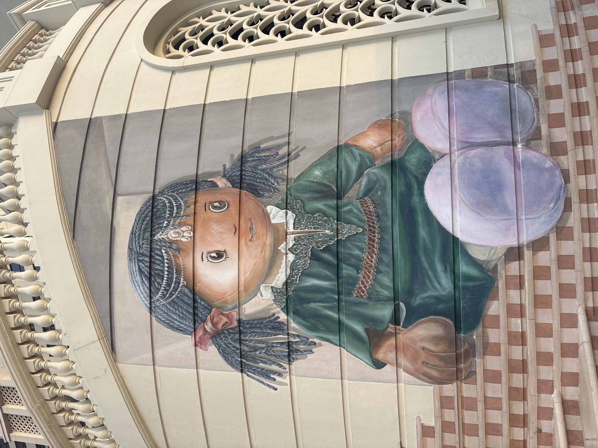 A mural of a doll is painted on a building's facade in Dubai, United Arab Emirates. The doll is depicted sitting on stairs, wearing a green dress adorned with intricate lace and a brown belt. The doll has braided dark hair with pink ribbons and a bejeweled headband. The building features architectural elements such as a balustrade and an ornate window. The mural is painted on a wall with horizontal paneling.