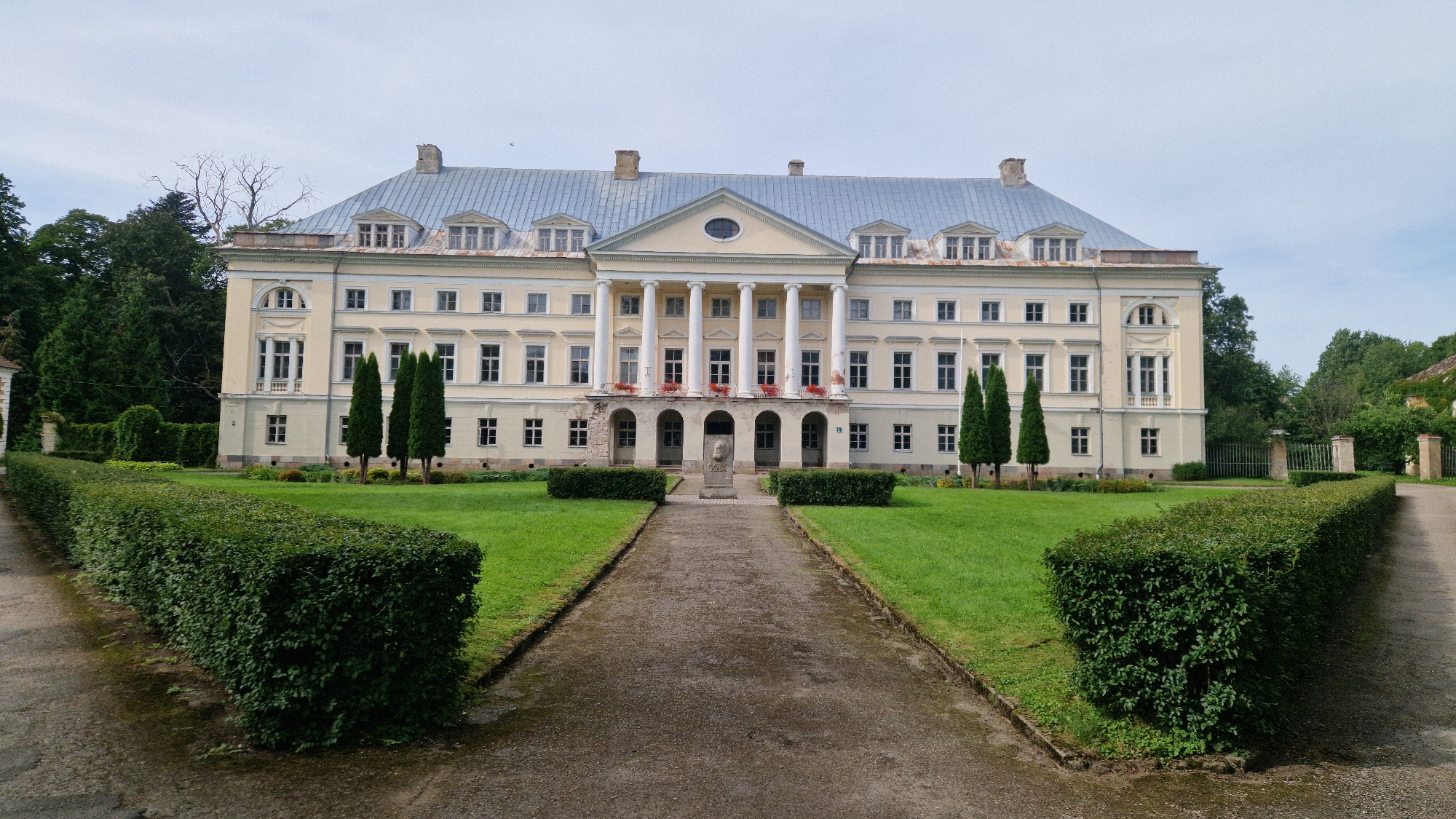 This is a full, eye-level, outdoor shot of Kazdanga Palace in Kazdanga, Latvia, on an overcast day. The pale yellow, neoclassical palace dominates the center of the frame, featuring a central portico with six large columns, a triangular pediment with an oval window, and a symmetrical facade with numerous windows and dormers. A gravel pathway leads from the foreground to the palace entrance, flanked by manicured green lawns and hedges. Two tall, slender evergreen trees stand to the right of the pathway and in front of the palace. In the center of the pathway, a stone pedestal with a bust of a person is visible. The palace grounds transition to dense green trees on the left and right sides of the frame, with a fence and more trees visible in the distance on the right. There are no people or animals visible in the image. The sky is a pale, hazy grey.