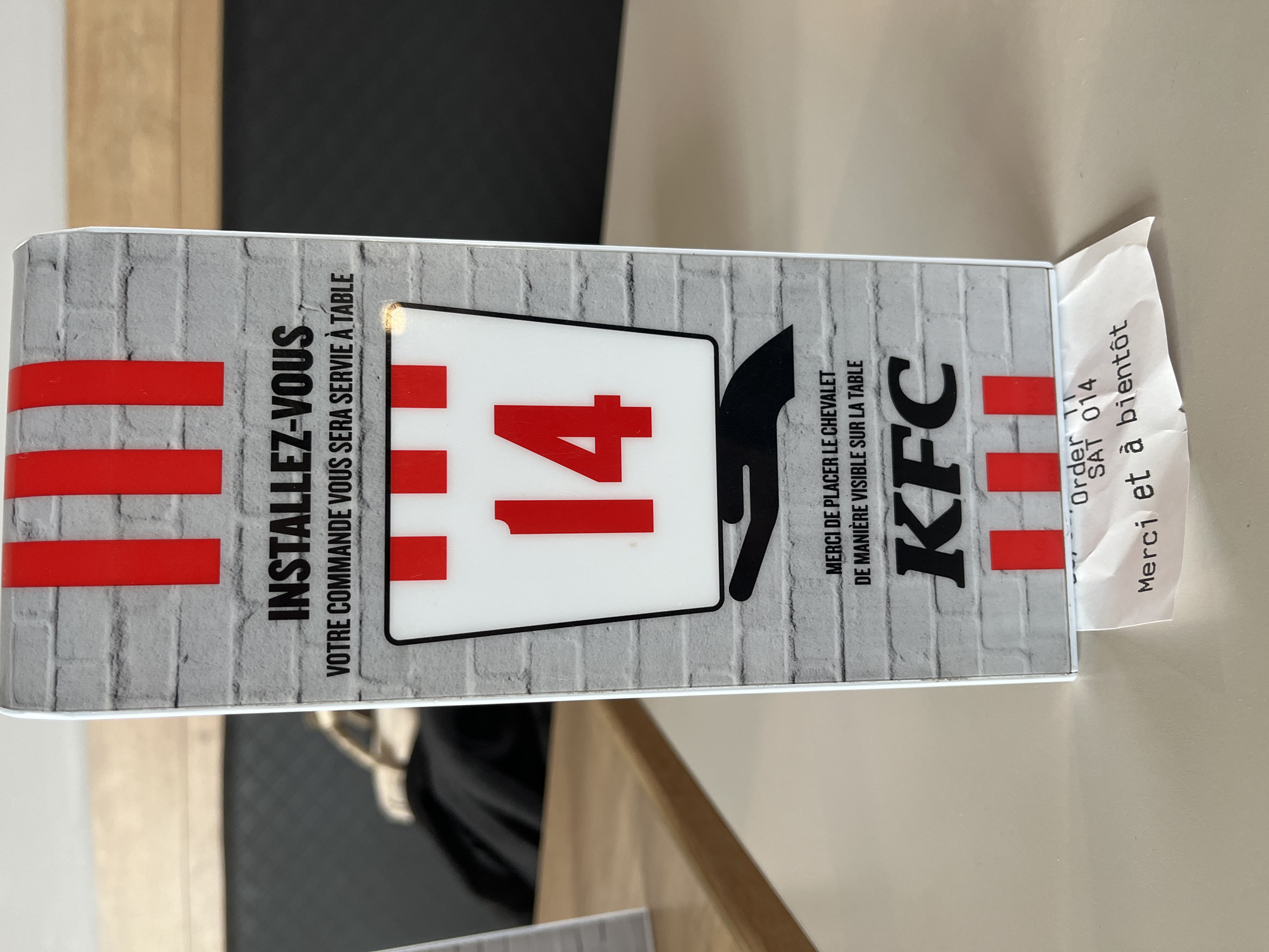 The image shows a table tent at a KFC restaurant. The tent is printed with a faux brick wall pattern and features the KFC logo, the number "14," and the phrase "INSTALLEZ-VOUS VOTRE COMMANDE VOUS SERA SERVIE À TABLE," which translates to "Make yourself comfortable, your order will be served at the table." It also includes the text "MERCI DE PLACER LE CHEVALET DE MANIÈRE VISIBLE SUR LA TABLE," which translates to "Please place the tent visibly on the table." A receipt is protruding from the side of the tent, with the order number "014" and the phrase "Merci et à bientôt," which translates to "Thank you and see you soon." The restaurant is located in Andrézieux-Bouthéon, France.
