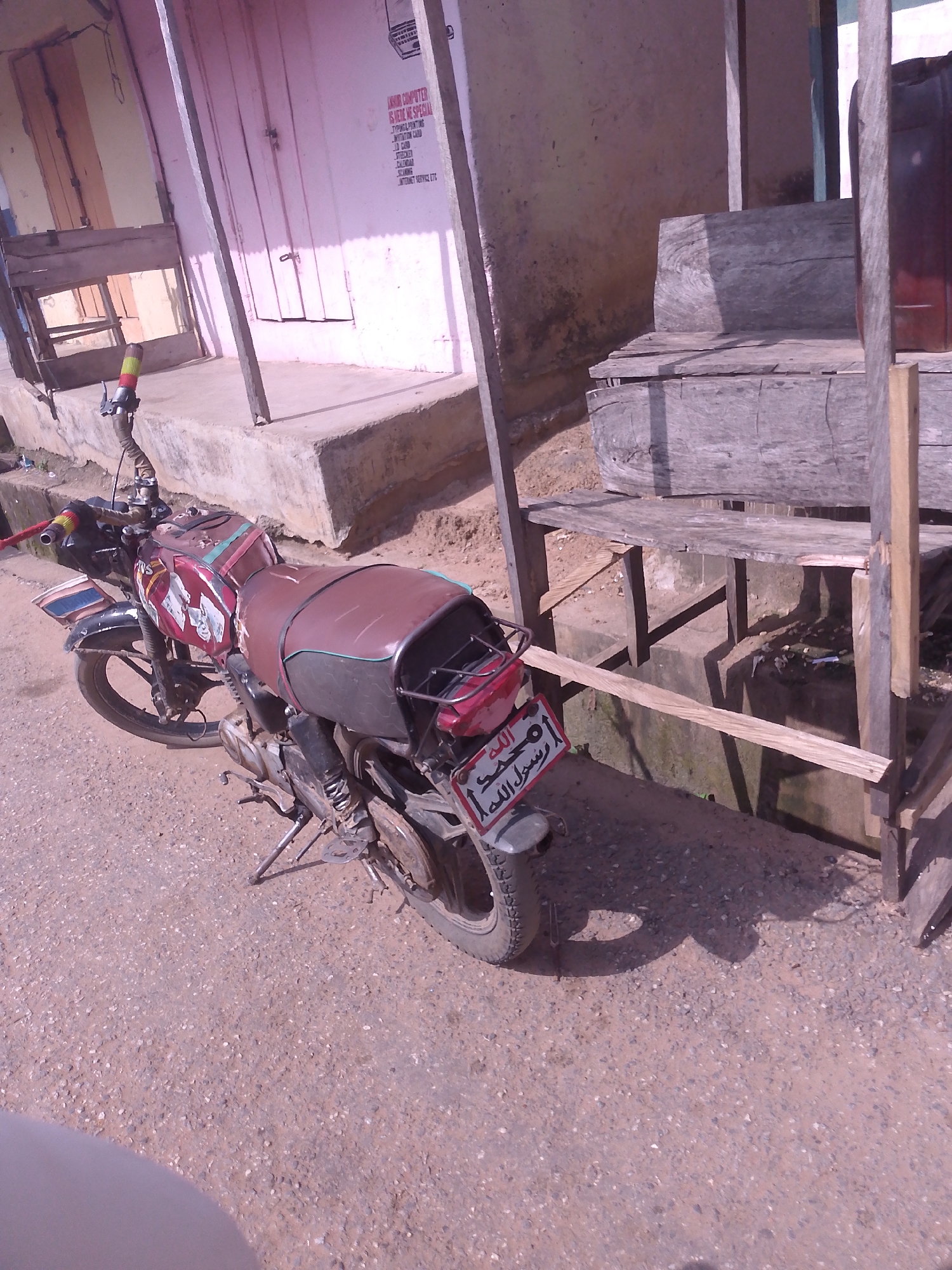 The image shows a street scene in Serti, Nigeria, with a parked motorcycle as the central subject. The motorcycle, a red and brown model, is positioned on a rough, gravelly surface. It appears to be a common, utilitarian model. A license plate with Arabic script is visible on the rear of the motorcycle. In the background, a building with a pink facade and wooden doors is present. The building's wall displays some text, possibly a business advertisement. Adjacent to the building, a wooden structure, possibly a stall or shelter, is erected with wooden planks and supports. The overall environment suggests a commercial or residential area within the city. No people are visible in the frame.