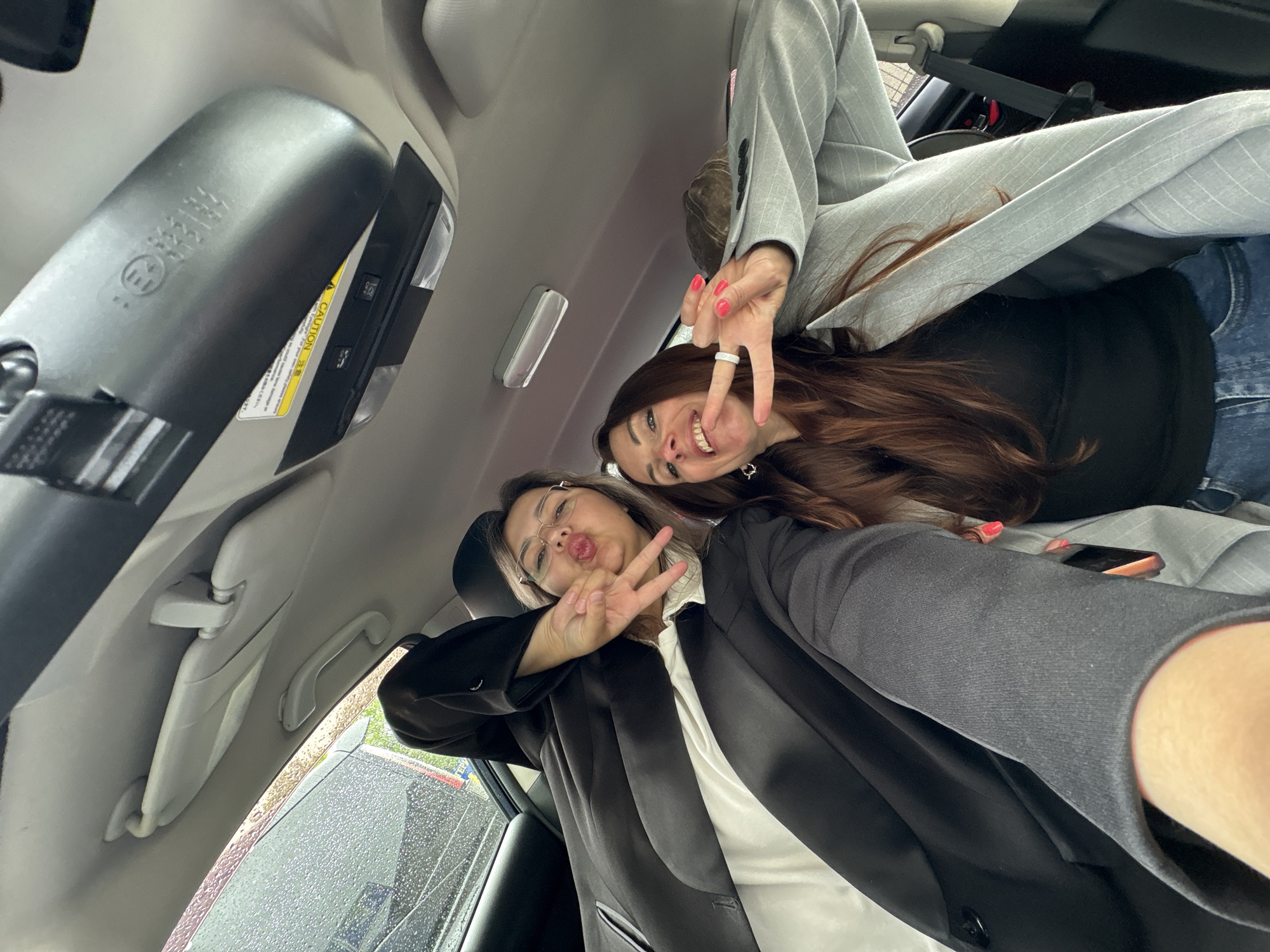 Two young women are captured in a playful selfie taken inside a car, likely in Birobidzhan, Russia. The image is taken from a low angle, looking upwards towards the car's ceiling, and appears to be inverted.
The woman in the foreground, wearing glasses and a black blazer over a white top, puckers her lips while flashing a 'peace' sign with both hands. Her light brown hair frames her face. Behind her, the second woman with long reddish-brown hair smiles broadly at the camera, also making a 'peace' sign with her right hand. She wears a grey pinstripe suit jacket, blue jeans, and has bright pink nail polish on her fingers, along with a white ring on her right ring finger.
The car's interior is visible, including the light-colored roof lining, sun visors, and a grab handle. An overhead console is prominent, featuring lights and buttons. On this console, a yellow "CAUTION" label with fine print is partially visible. Another label on the console displays what appears to be a stylized 'E' logo and some blurry, inverted text that might be East Asian characters. A button labeled "SOS" is also discernable on the console.
Through the car windows, rain or water droplets are visible, indicating wet weather outside. The overall mood of the image is cheerful and casual, with both subjects expressing joy.